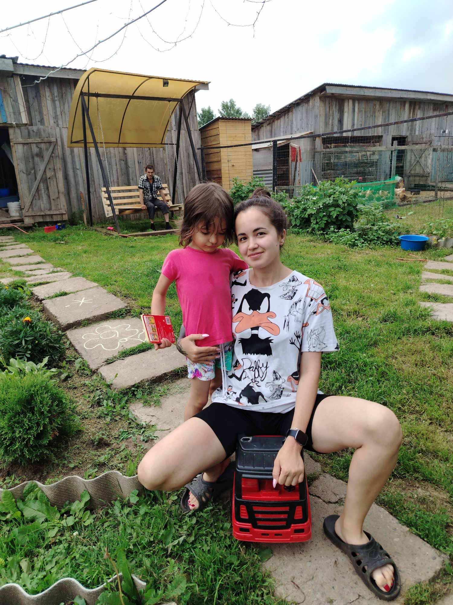 In the city of Gozhan, Russia, a woman is kneeling on a concrete pathway in a grassy yard, holding a red toy truck. She is wearing a white t-shirt with cartoon characters and black shorts. A young girl, wearing a pink t-shirt and patterned shorts, stands beside her, holding a small red book. In the background, a man sits on a wooden swing set beneath a yellow canopy, next to a rustic wooden building. Other structures, including a chicken coop, are visible further in the distance. The environment suggests a rural or semi-rural setting, likely a backyard or a small farm.
