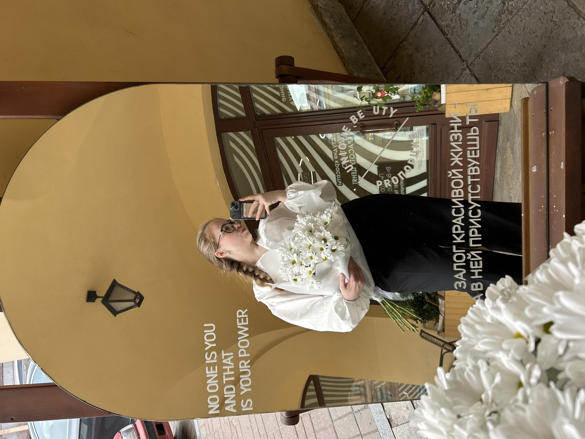 A woman takes a mirror selfie while holding a bouquet of white chrysanthemums. The mirror has text in English "NO ONE IS YOU AND THAT IS YOUR POWER" and Russian. The reflection includes a door with striped awnings and the word "BEAUTY" with a partial business name "Порция" visible. The scene is in Saint Petersburg, Russia.