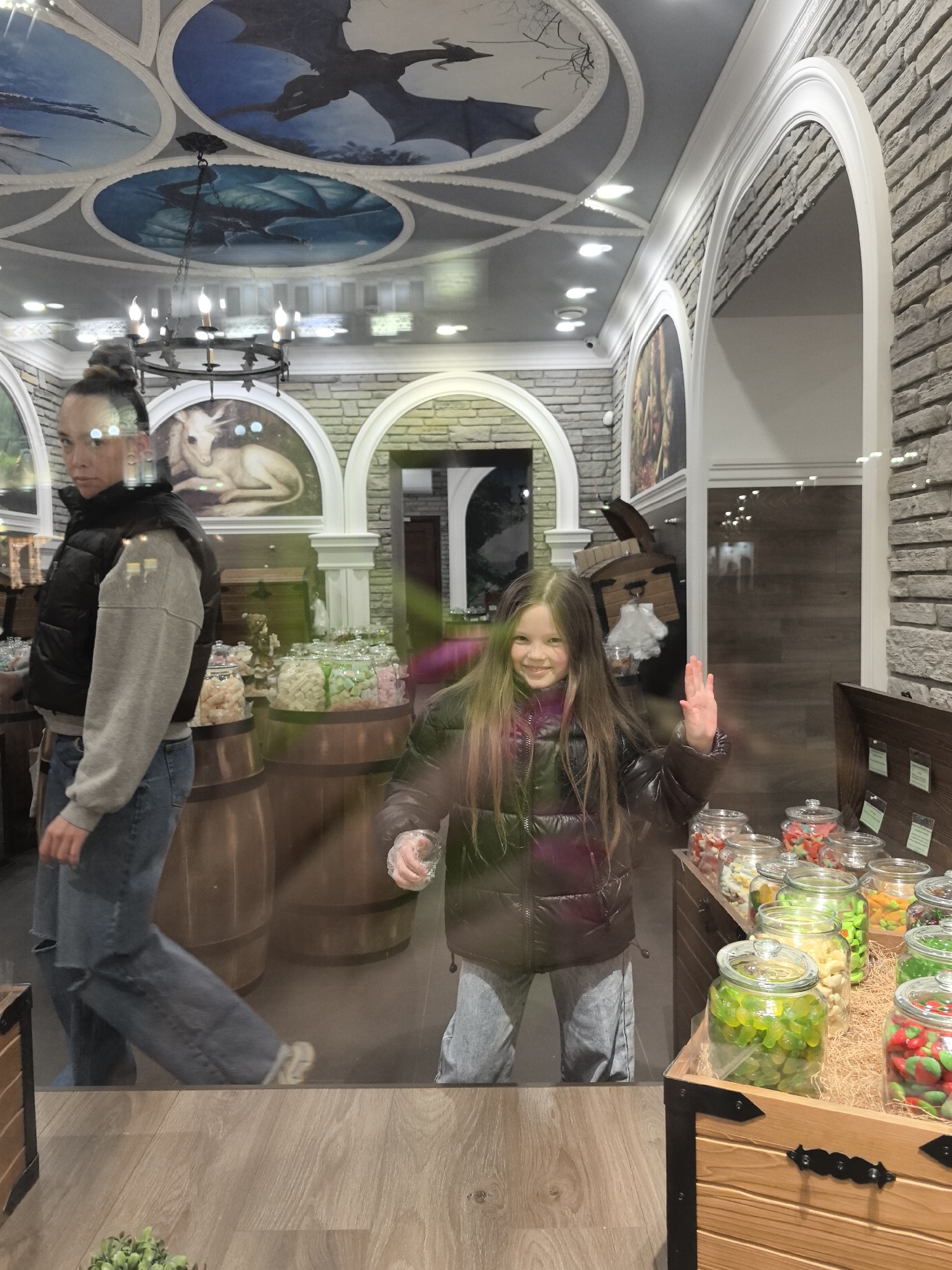 Captured from a point outside a shop in Nizhny Novgorod, Russia, the photograph offers a view into a confectionery. A young girl, donned in a dark puffer jacket and jeans, extends her right hand in a wave towards the viewer. A woman with her hair in a bun, dressed in a black vest over a grey sweater and jeans, walks away from the camera and to the left. The interior features candy in jars, arranged on a wooden display resembling a treasure chest. Barrels topped with candies are also visible. The ceiling is decorated with dragon and unicorn motifs, and a chandelier hangs near the shop window. The shop's architecture includes arched entryways and stone brick walls.