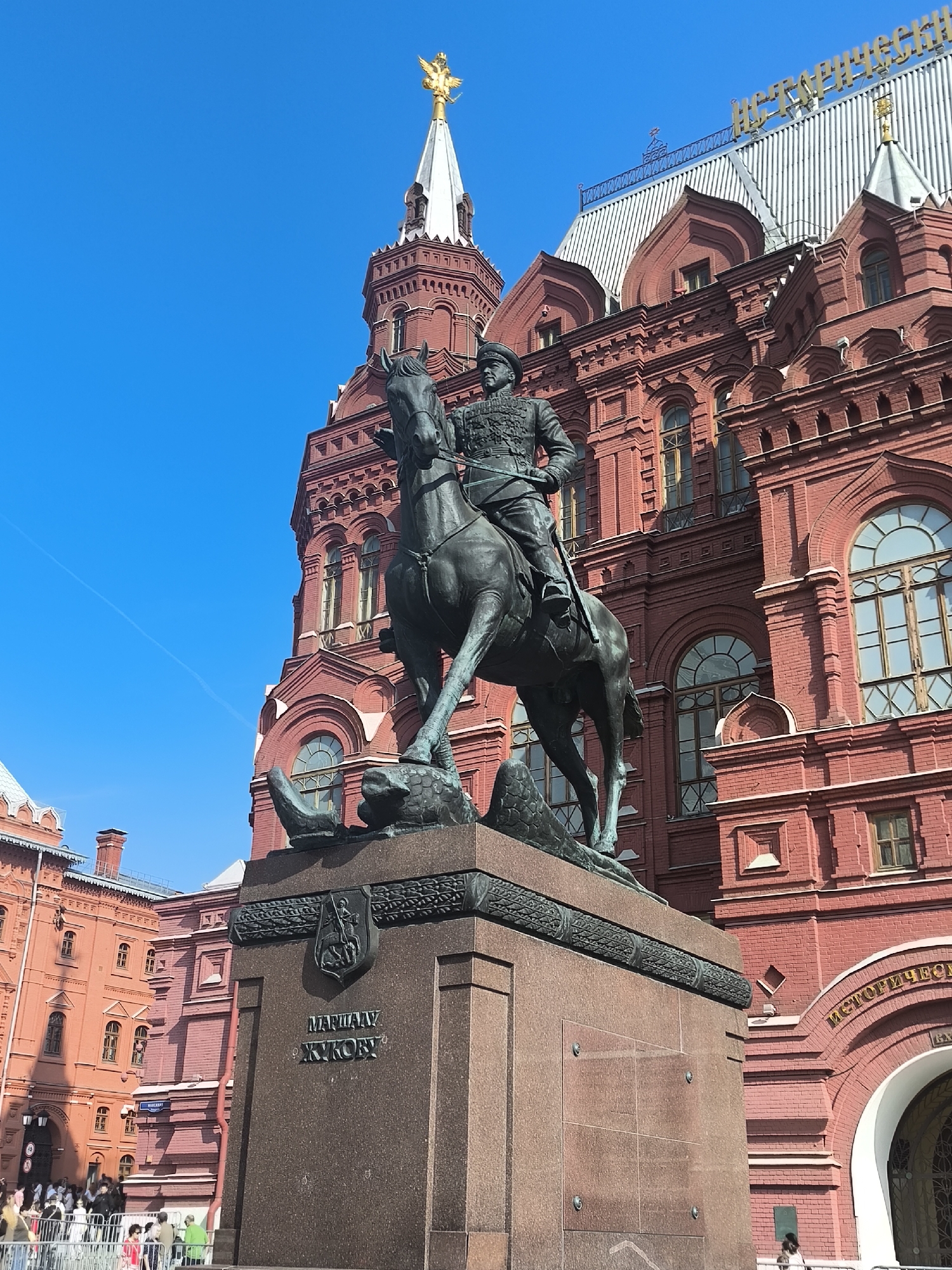 The image captures a statue of Marshal Zhukov on horseback in front of the State Historical Museum in Moscow, Russia. The bronze equestrian statue dominates the foreground, with the ornate, red-brick facade of the museum in the background. The sky is a clear, bright blue. In the lower-left corner, a crowd of people can be seen, suggesting the area is a public space.