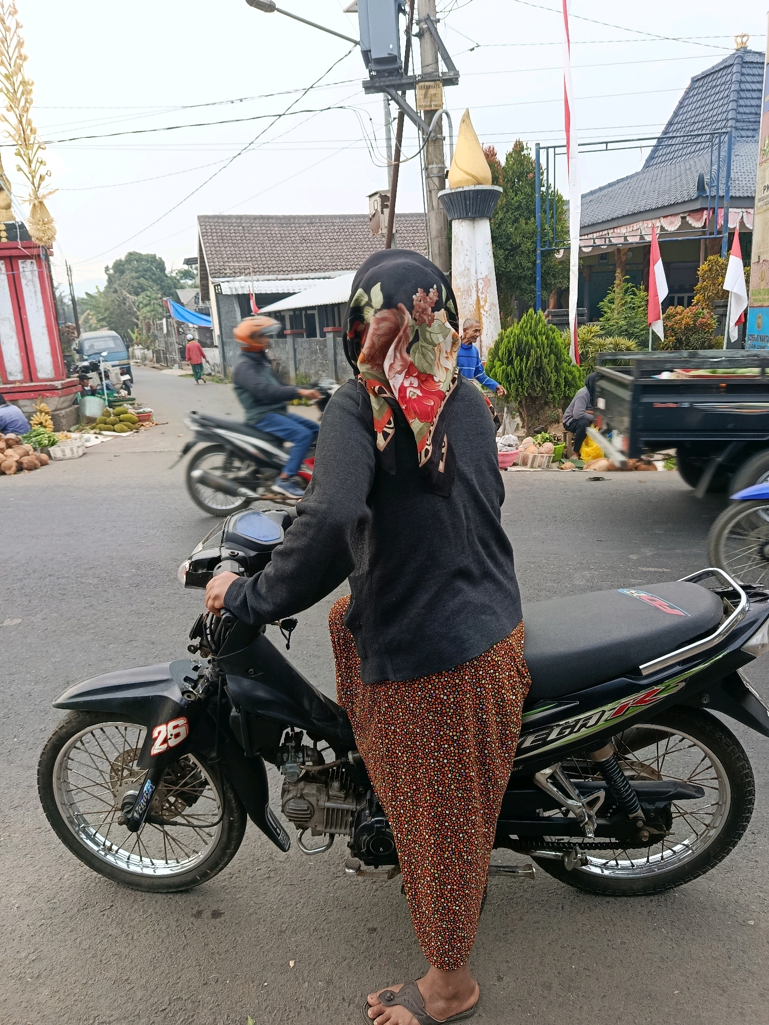 In Tajinan, Indonesia, a person is seated on a black motorcycle, their back to the camera. The person wears a dark gray long-sleeved shirt and a patterned headscarf with red, green, and white floral motifs. Their pants are also patterned with small red, orange, and white dots on a dark background. The motorcycle is a black street bike with a visible engine and chrome accents. A license plate with the number "25" is visible on the front fender. The motorcycle's seat is black with a colorful decal. The scene is set on an asphalt road, with other vehicles and buildings in the background. To the left, a vendor is selling fruits, including coconuts and durians. A blue van is parked behind the vendor. In the distance, a few houses with tiled roofs and flags are visible. The sky is overcast.