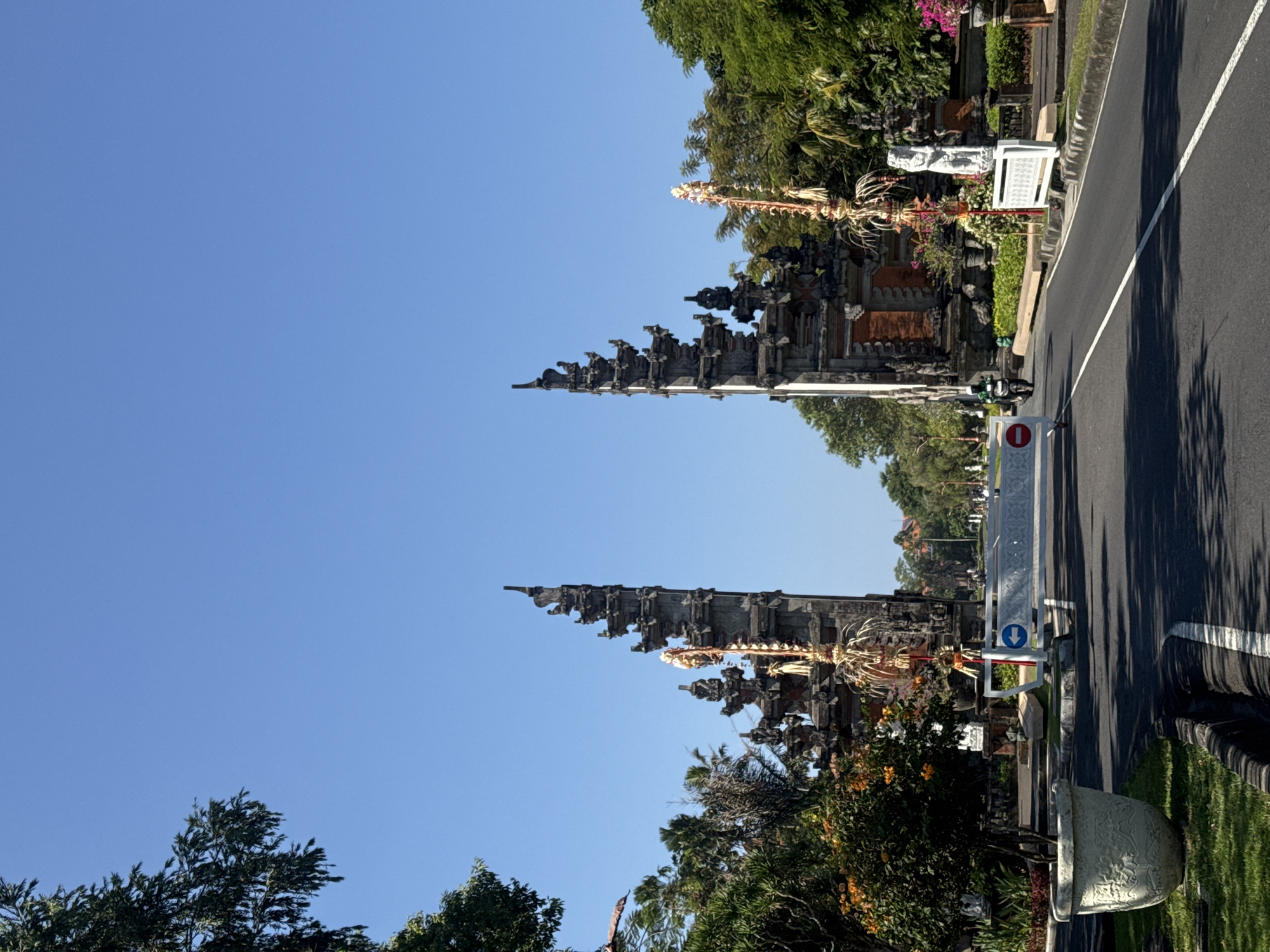 A vertical, eye-level, wide shot captures a grand Balinese temple gate in Kuta Selatan, Indonesia, under a clear blue sky. The intricately carved stone gateway, a prominent feature, is flanked by two towers, each adorned with traditional Balinese motifs. The road leading through the gate is paved with asphalt and marked with white lines.
On the left side of the road, a large, ornate concrete planter holds a small tree with orange flowers. The right side of the road features lush green trees and decorative plants, including a white statue of a person. In the foreground, a white barrier with red and blue signs indicates a temporary road closure or traffic direction.
A lone motorcyclist in a green jacket is seen riding through the gateway on the right side of the road. The architectural style of the temple gate suggests a traditional Balinese Hindu influence. The overall scene depicts a sunny day in a tropical location, with elements of both cultural heritage and modern infrastructure.
