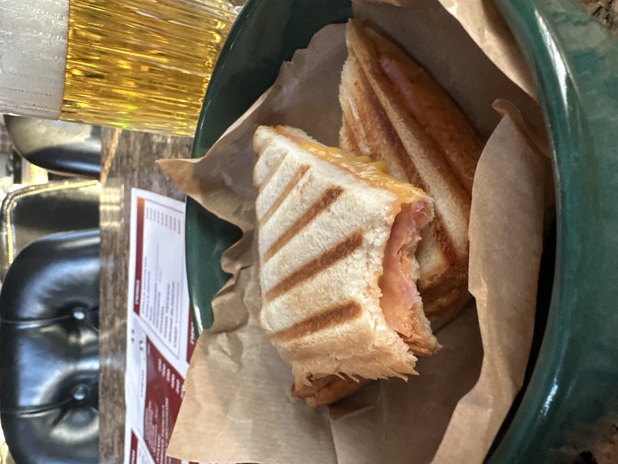 The image shows a partially eaten grilled sandwich, cut in half, and placed in a green bowl lined with brown paper. The sandwich appears to contain ham and cheese. A menu card with red and white text is visible next to the bowl. Parts of a black chair and a shiny, gold-colored object are also in the frame. There is no information to indicate that the location is in Votkinsk, Russia.