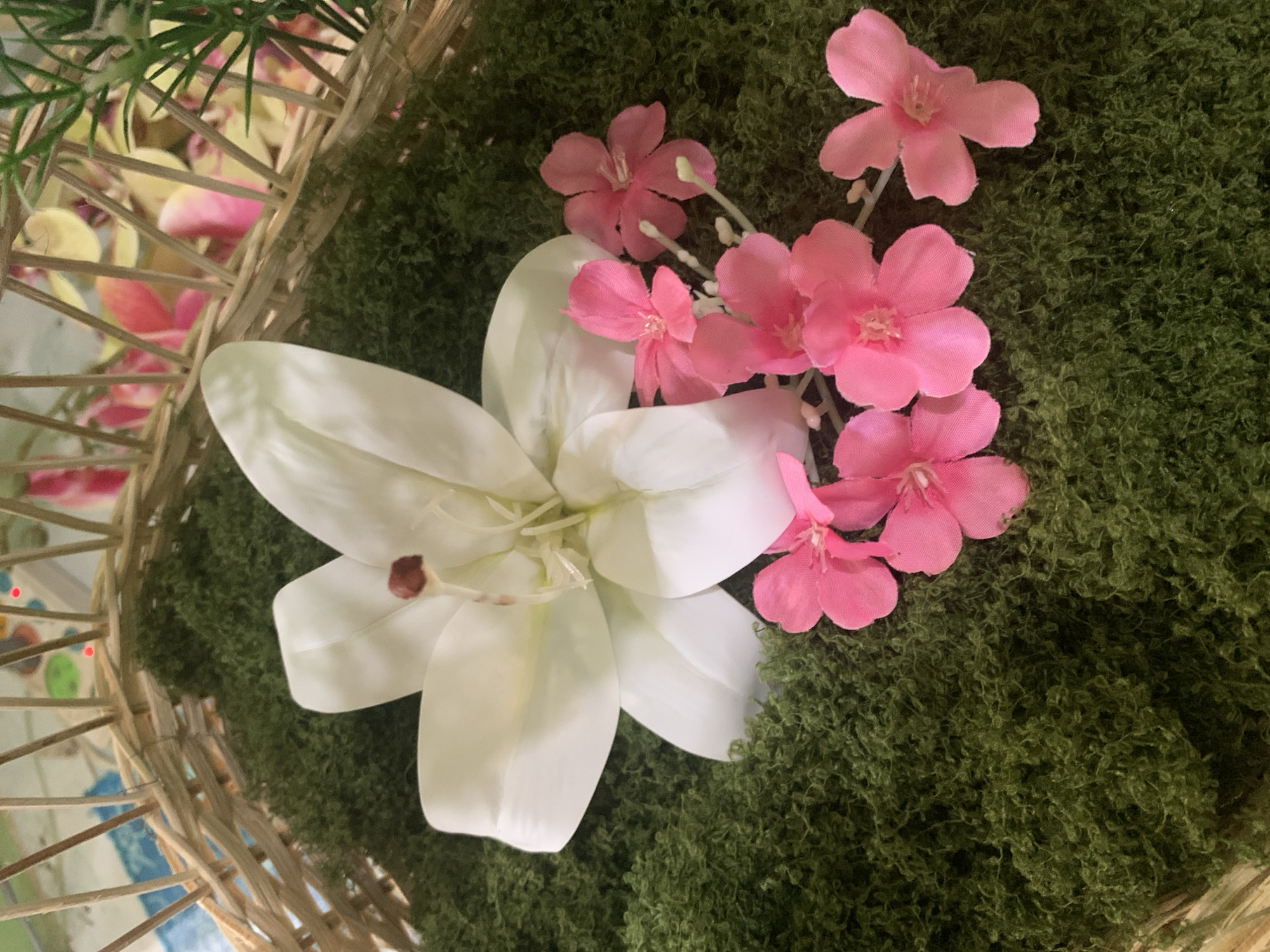 A close-up shot shows a white lily and several small pink flowers nestled on a bed of green artificial moss, arranged within a rustic woven basket. The basket's woven texture is visible, with some of its supports reaching upwards. In the background, out of focus, are colorful elements, possibly more flowers and greenery, suggesting an outdoor or decorative setting. The lighting is soft, casting gentle shadows and highlighting the delicate details of the artificial flora. The scene does not depict any people or specific actions, focusing purely on the floral arrangement. The location in Kuala Pembuang, Indonesia, is not visually discernible from the image itself.