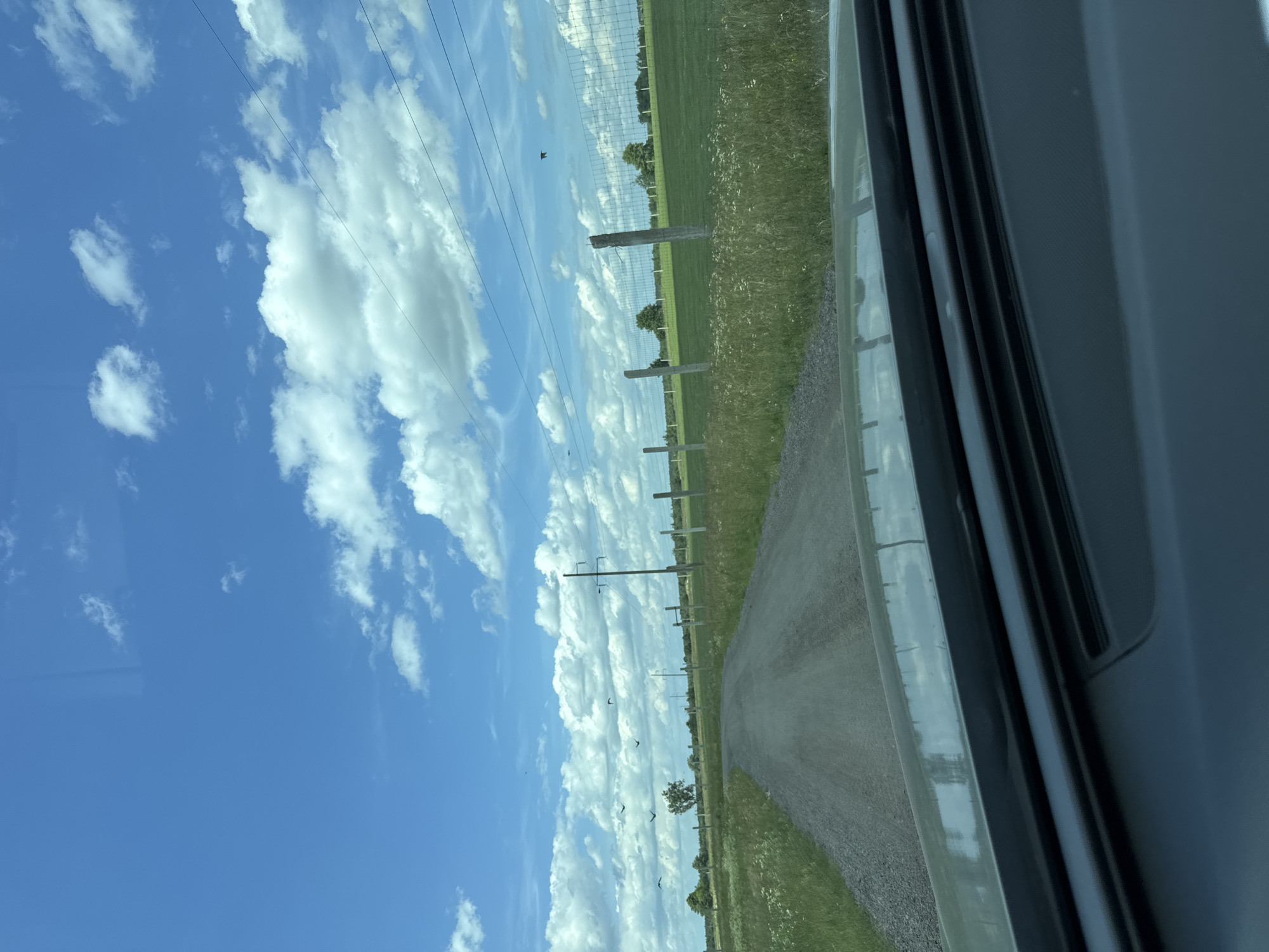 This media file depicts a rural scene in Orlivka, Ukraine, from the perspective of a vehicle. The image is oriented vertically, with the sky occupying the majority of the frame on the left and the ground and a dirt road on the right.
The sky is a vibrant blue, dotted with fluffy white clouds, suggesting a clear and pleasant day. A few power lines are visible stretching across the sky, supported by utility poles that are interspersed along the side of the road. These poles are accompanied by wooden fence posts, indicating a boundary or enclosure.
The road itself appears to be unpaved, made of gravel, and curves gently into the distance. To the right of the road, a field of green grass and wild flowers stretches out, bordered by a fence. In the background, several trees are scattered across the landscape, and the overall impression is one of open countryside.
There are no people visible in the image. The activity taking place is implied to be a journey or drive along this rural road. Notable details include the presence of the power lines and fence posts, which are common features in rural Ukrainian landscapes. The time of day appears to be daytime, likely late morning or afternoon, given the brightness of the sky and the quality of the light. The weather seems fair, with a mix of sun and clouds.
The composition, looking out from within a vehicle, gives a sense of movement and observation. The dark area on the far right of the image appears to be the interior of the vehicle, possibly a window frame or dashboard. There is no visible text in the media file.