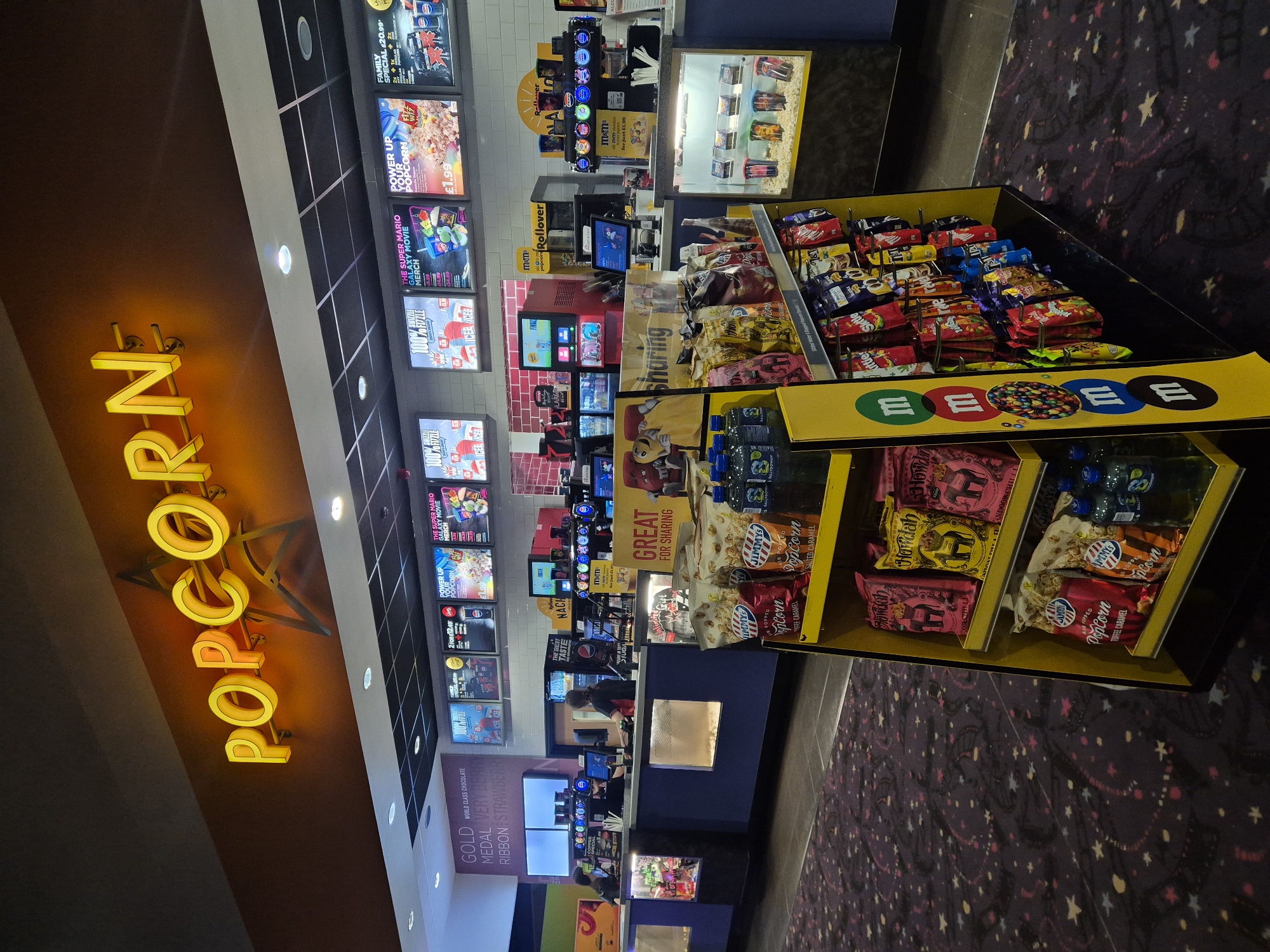 This photograph captures the interior of a concession area at a cinema, potentially in West Suffolk, United Kingdom, as it showcases products and signage commonly found at movie theaters. The scene is organized with various displays and points of sale.
The main subjects in the image are the product displays featuring snacks and drinks. Popcorn is advertised prominently with a large, illuminated "POPCORN" sign, which is the most eye-catching element. Movie posters and advertising boards can be seen lining the upper wall, promoting current films and special offers.
In the foreground, there's a display filled with sweets, including boxes of M&Ms and assorted chocolate bars. Below, another display level presents bags of popcorn and bottled water. These items are arranged for easy customer access.
Behind the displays, there is a counter area where a cinema employee may be stationed, although their specific actions are not visible in detail. Additional screens show digital advertisements or point-of-sale information.
The environment has a commercial feel, with the space designed to facilitate the quick purchase of snacks and drinks before a movie. The flooring has a dark, star-patterned carpet. The image showcases the typical atmosphere and layout of a cinema's concession stand.