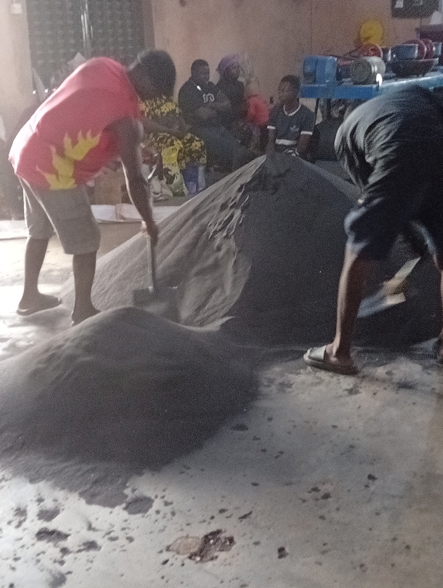 Gura Top, Nigeria: Two men are working with large piles of dark sand on a concrete floor. One man, wearing a red shirt with yellow flames, khaki shorts, and sandals, uses a shovel to move sand. The other man, wearing a dark shirt, dark shorts, and sandals, is also working with the sand. In the background, other individuals are seated, including one wearing a dark shirt with a white emblem. A blue machine is visible in the background.