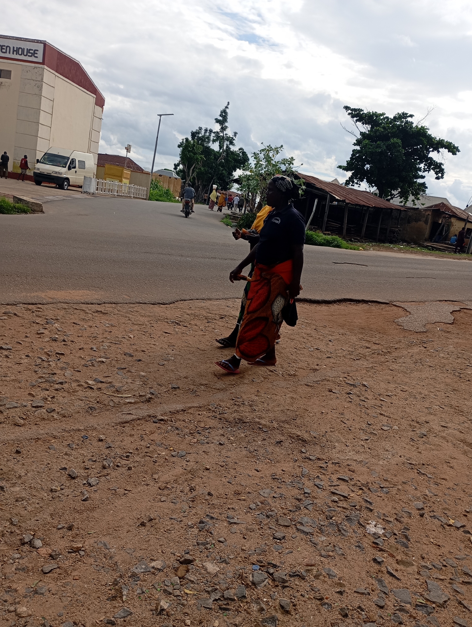 In Shendam, Nigeria, on a cloudy day, a paved road is visible, with a dirt shoulder on the left. A white van is parked on the left side of the road, in front of a building with "EVEN HOUSE" signage. Several people are walking on the road and sidewalk, including a woman in a dark blue shirt and a colorful patterned skirt, who is walking towards the right side of the image. She is accompanied by another person in a yellow shirt. Further down the road, a motorcyclist is visible. On the right side of the road, there are some dilapidated structures with corrugated metal roofs. The ground in the foreground is covered with reddish-brown dirt and scattered stones.