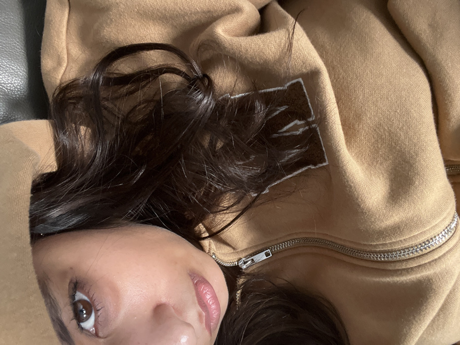 A close-up image features a person with dark brown hair lying down, with their face partially visible. The focus is on their eye, nose, and lips. They are wearing a beige zip-up hoodie, with the zipper partially open. The hoodie features a dark brown patch with light-colored stitching. The texture of the fabric is visible, and the overall lighting creates shadows and highlights. The background appears to be a dark-colored surface.