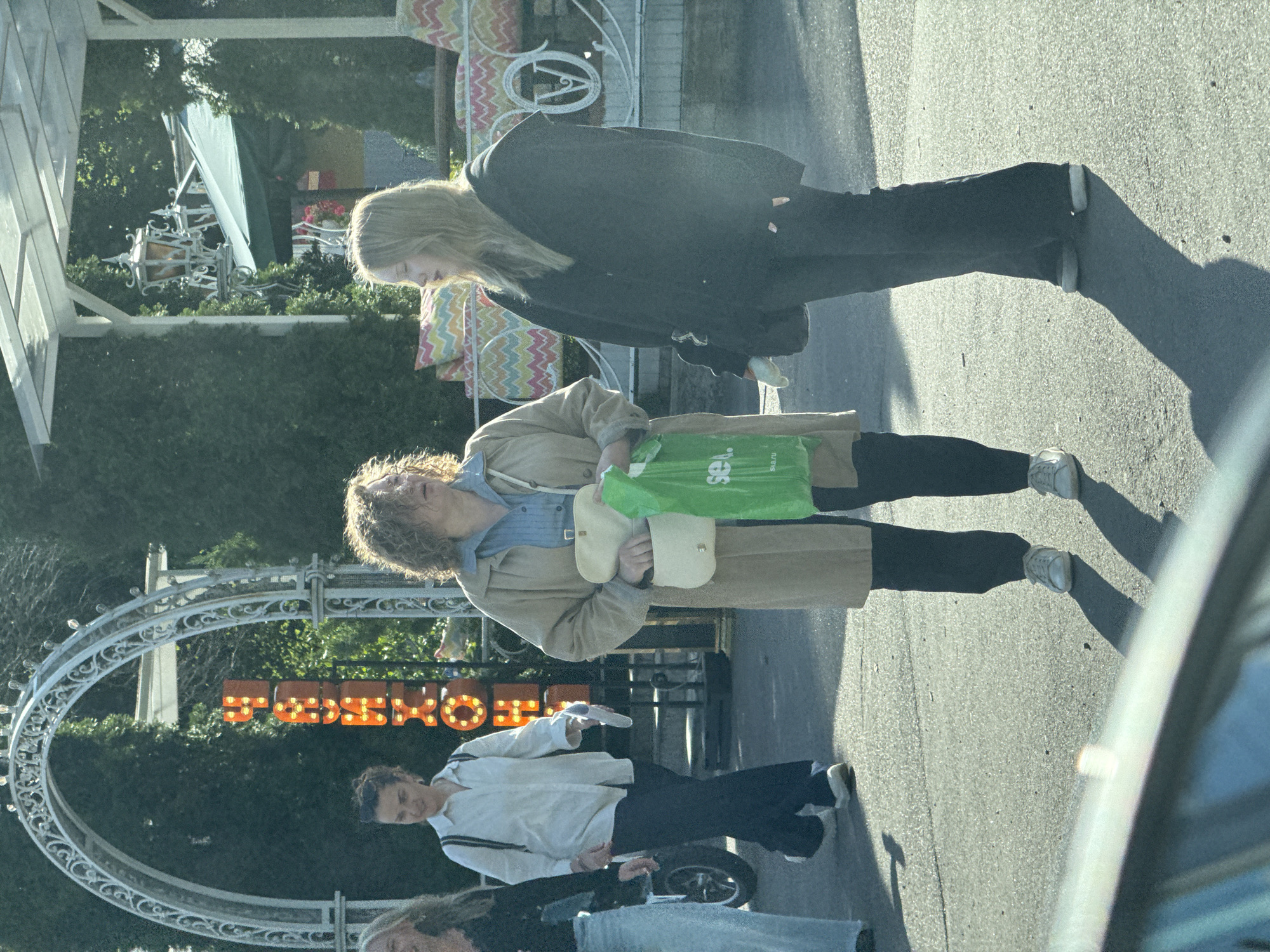 Four individuals are captured in a medium shot on a sunlit street in Saint Petersburg, Russia. The scene appears to be near an amusement park or entertainment venue, indicated by a decorative archway with lights and a vertical neon sign with orange letters spelling "RUSHOUX" in Russian, and beyond that, a green awning with colorful cushions on display. The pavement is grey and textured, with strong shadows cast by the subjects and objects.
On the left, two women stand near a motorcycle. The woman in the foreground has long, light brown hair and is wearing a black top and light blue jeans. She is looking slightly to her right. Behind her and to the left, a woman with dark, curly hair is wearing a white button-down shirt and dark pants, carrying a dark backpack. She is looking forward and to her left.
In the center and to the right, two women stand closer to the foreground. The woman on the left, closer to the center, has curly, brown hair and is wearing a light brown trench coat over a blue shirt and dark pants. She holds a light-colored purse in her left hand and a bright green plastic shopping bag with a logo that appears to read "sea.ru" in her right hand. She is looking forward. To her right, a woman with long, blonde hair wears a dark, oversized hooded jacket and dark pants. She is looking forward and slightly downwards, with her right hand resting on her hip.
The overall lighting suggests it is a bright, sunny day. The background features dense green trees and architectural elements of what appears to be an amusement park or similar recreational area. The composition is dynamic, with the subjects positioned at varying distances and facing different directions.