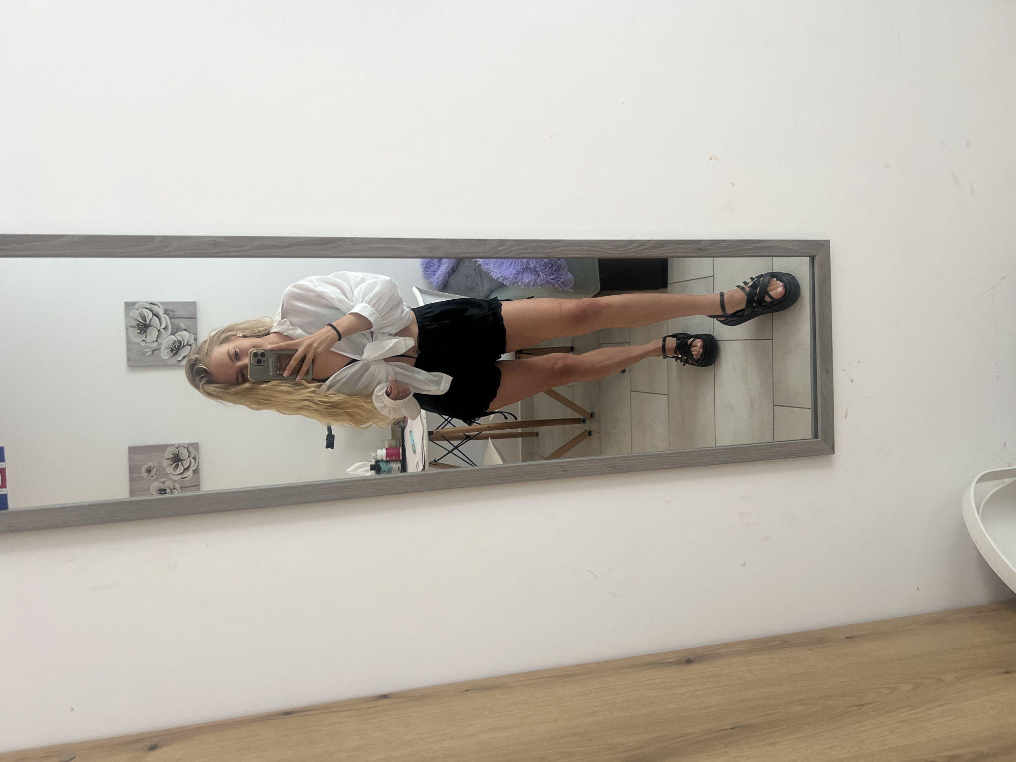 A young woman with long blonde hair, wearing a white long-sleeved shirt and black shorts, stands in front of a large rectangular mirror with a grey frame, taking a selfie with her smartphone. She is wearing black platform sandals on her feet. Her face is partially visible as she holds the phone up to capture her reflection.
The mirror reflects a room with light-colored walls and tiled flooring. Behind the woman's reflection, a light purple fluffy blanket is draped over what appears to be a bed or couch. To her left (in the reflection), two small square framed pictures featuring white floral designs are mounted on the wall. A wooden stool or small table with an X-shaped frame is also visible in the reflection. To the right of the reflected woman, there's a small collection of bottles or containers on a surface. Outside the mirror's reflection, the foreground features a wooden surface, and the wall is white with some faint marks. No discernible text is visible. The scene appears to be an indoor setting, possibly a bedroom or dressing area.