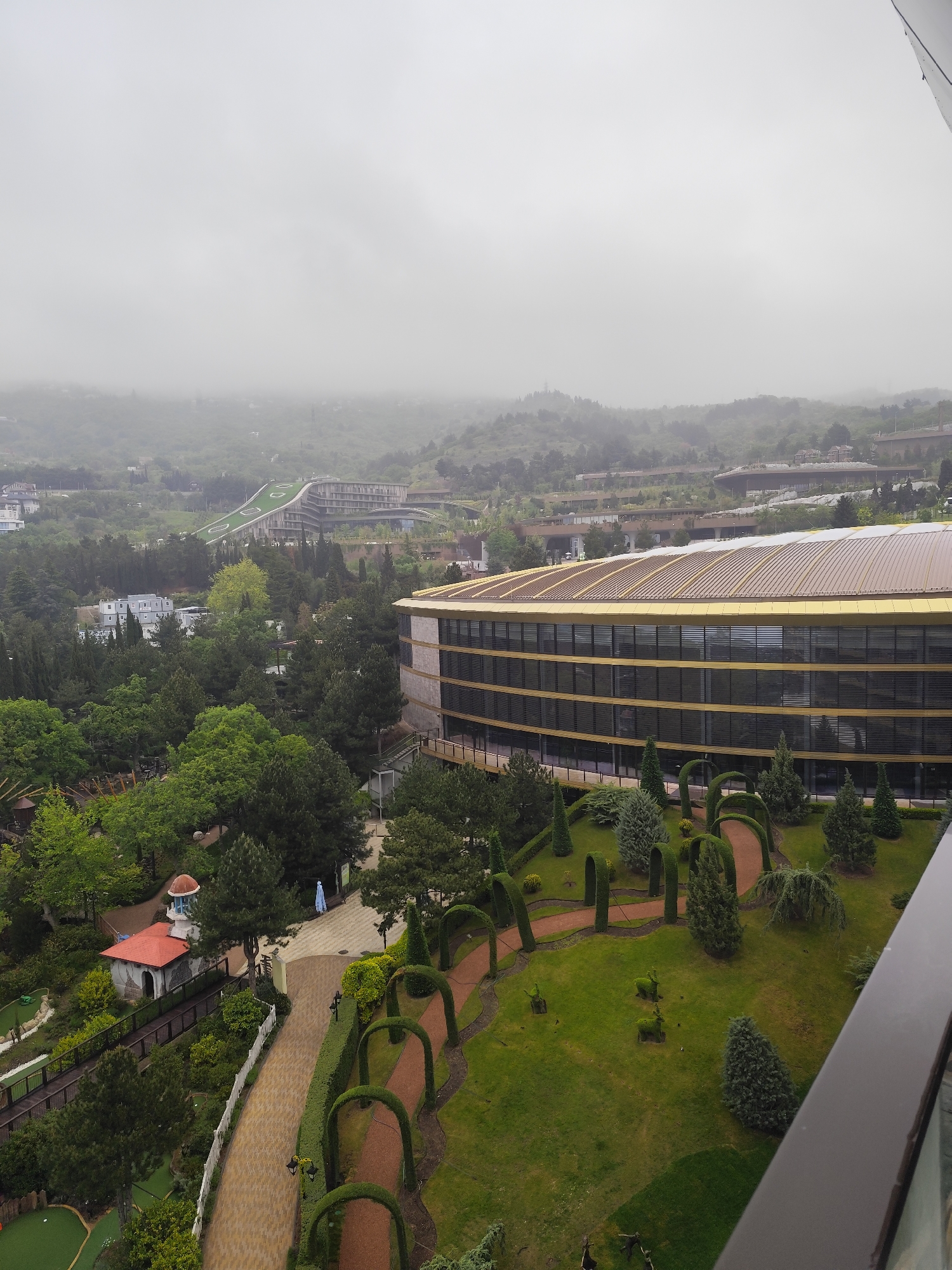 A high-angle, overcast daytime shot shows a sprawling landscape in the urban district of Симеизский поселковый совет, Ukraine.
Dominating the midground is a large, contemporary building with a distinctive curved design and a golden roof. The building's facade features multiple tiers of dark-tinted windows. Adjacent to the building is a meticulously maintained garden area with shaped hedges and pathways.
To the left of the building, a pathway leads away from the camera, passing by a small building with a red roof. Beyond the pathway, a lush green area is visible.
In the background, a mountainous landscape is partially obscured by a heavy layer of mist or fog. Another building, which may also be a hotel, is visible in the background.