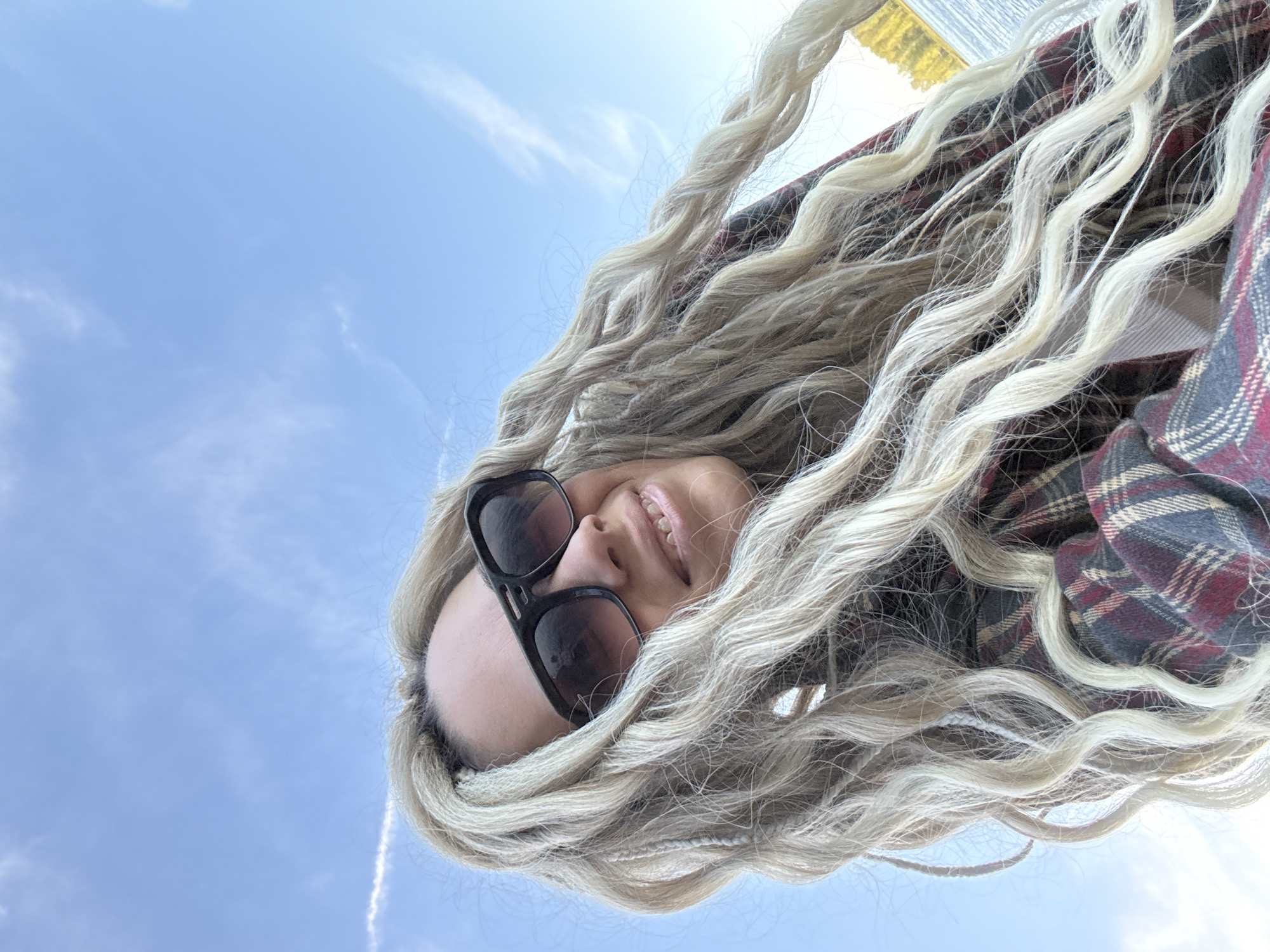 A woman with blonde, braided hair is smiling into the camera, which is angled upward. She is wearing dark sunglasses and a red and blue plaid shirt. The background is a blue sky with a few wispy clouds and a contrail. A body of water is visible at the top of the frame, likely a lake or river. The photograph was taken outdoors during daylight hours. The location is presumably Rusanovo, Russia.