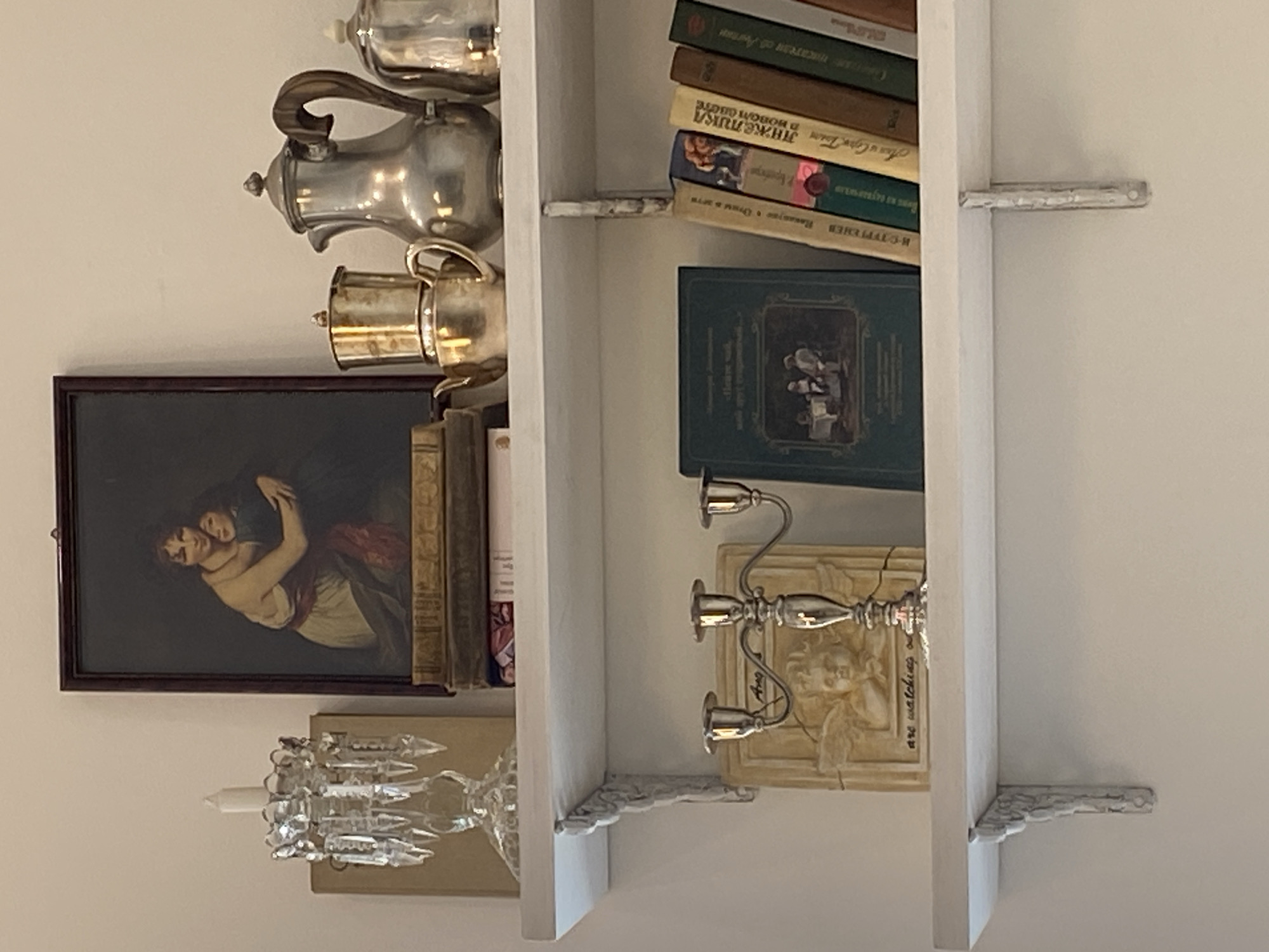 The image depicts a shelf display against a white wall. On the top shelf, a framed painting of two women embracing is centered, flanked by a crystal candelabra with a white candle to the left and stacked books and silver teapots to the right. The books are varied in size and color, with the teapots appearing to be a coffee pot and a larger teapot. On the lower shelf, a decorative relief of an angel with wings is positioned behind a silver candelabra with three arms. To the right of the candelabra, a stack of books is arranged vertically. The books on the lower shelf are varied in color, with some featuring illustrations and text in Cyrillic script. The overall scene suggests a curated display of decorative and literary items in an indoor setting.
