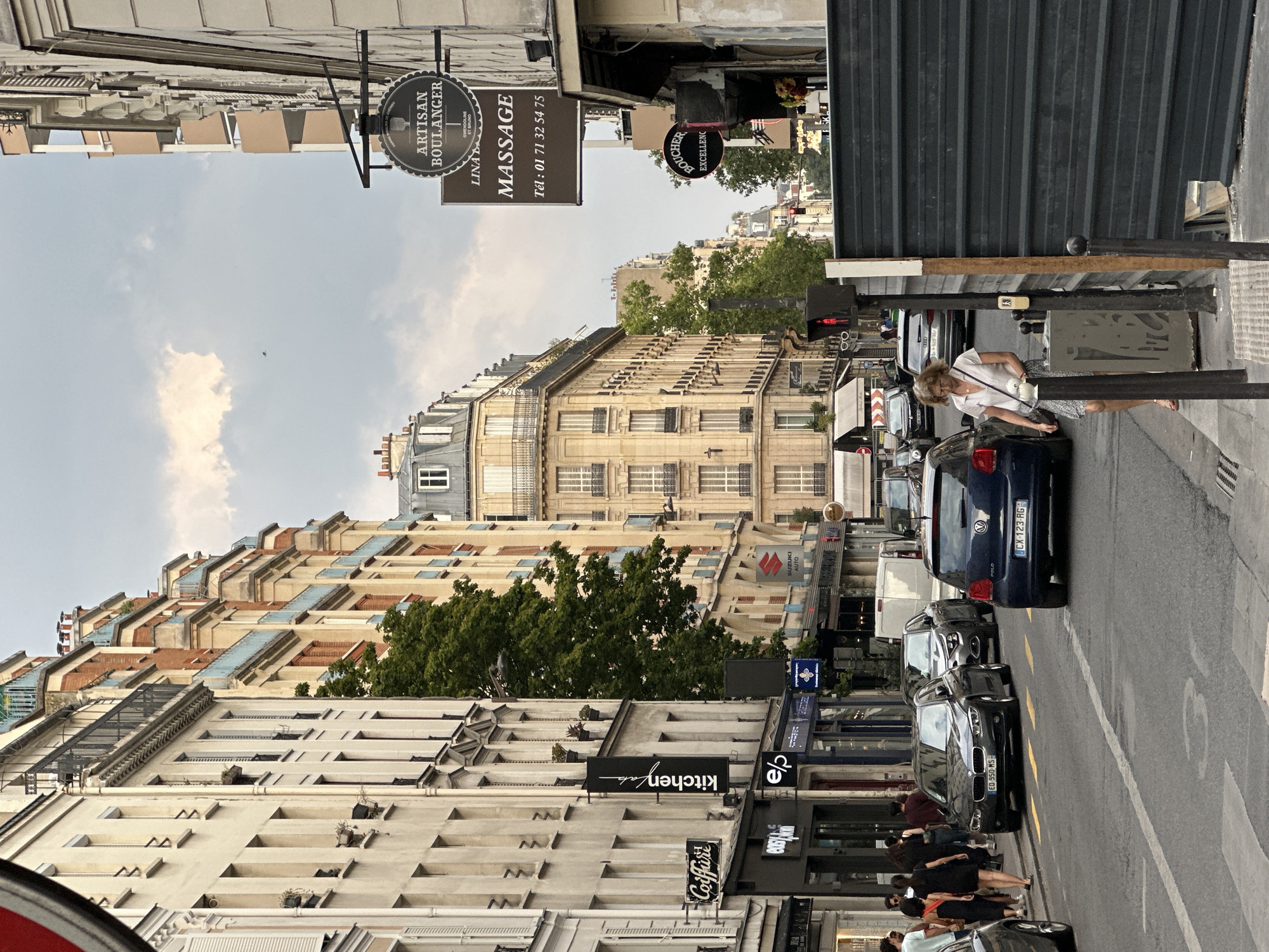 The image depicts a street scene in Paris, France, characterized by its classic Haussmannian architecture. Tall, cream-colored buildings with ornate facades, multiple floors, and balconies line the street. A prominent sign for "ARTISAN BOULANGER" hangs from one building, suggesting a bakery. Another sign advertises "MASSAGE" with a phone number.
The street itself is paved and appears to be a one-way thoroughfare with several cars driving or parked. Notable vehicles include a dark gray BMW sedan with the license plate "EO 950 MS," a blue Volkswagen Polo with license plate "CX 123 RG," and other sedans and SUVs. Pedestrians are visible on the sidewalk, including a woman in a light-colored shirt and patterned skirt walking towards the foreground. There are also several trees providing greenery amidst the urban landscape. The sky is partly cloudy, with hints of a bird in flight.
The scene suggests a typical day in a Parisian neighborhood, with shops, traffic, and people going about their daily activities. The presence of construction barriers on the right side of the image indicates ongoing work or renovations. The overall atmosphere is one of bustling city life within a historically rich architectural setting.