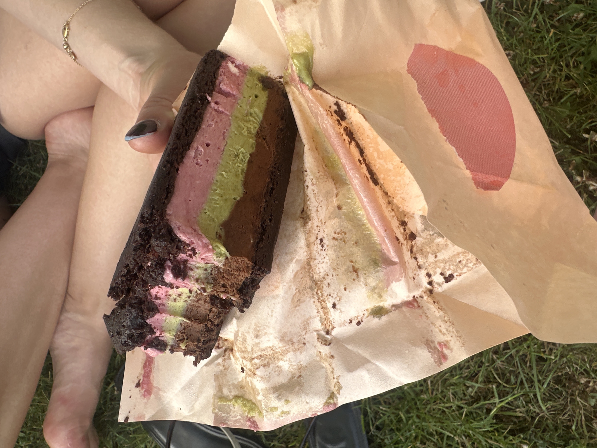 A person sits on grass in an outdoor setting, possibly in London, United Kingdom. The person holds a partially eaten rectangular dessert. The dessert has a dark chocolate crust, followed by pink, green, and dark brown layers. The dessert is held in a light brown paper bag, stained with remnants of the dessert. The background includes green grass. The person's legs are visible, along with a gold bracelet on their wrist. One fingernail is painted with a dark color with a light blue edge. Part of a shoe can be seen in the lower center of the image. A red patch is visible on the outside of the bag.