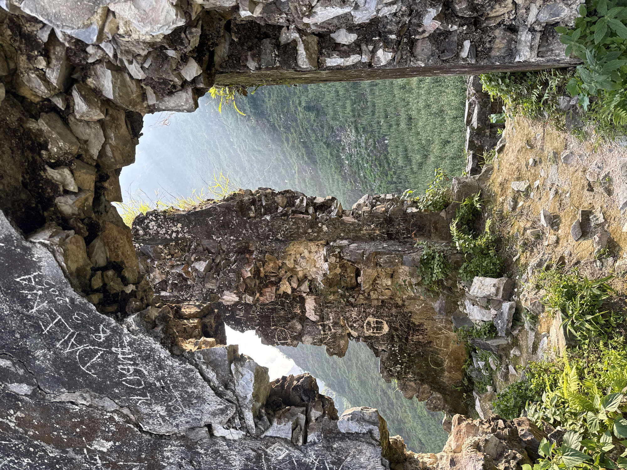 The image depicts a view through a ruined stone structure in Đường Thượng Commune, Vietnam. The structure is made of rough-cut stones and mortar, with openings worn or broken through the walls. Some stones are covered in graffiti. The view through the openings reveals a lush, green hillside obscured by mist. Vegetation grows on and around the stone structure.