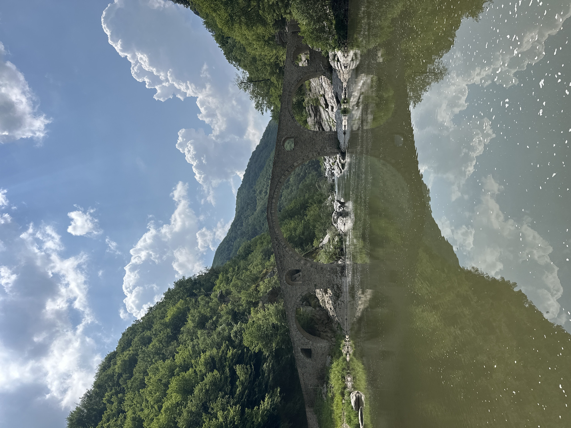 An upright-oriented, wide shot captures a picturesque natural landscape featuring an ancient stone arched bridge spanning a tranquil river in Diadovtsi, Bulgaria. The sky, occupying the upper portion of the frame, is a clear blue adorned with numerous fluffy white cumulus clouds. Below the sky, a steep, densely forested mountainside, vibrant with green foliage, dominates the left side of the scene.
The central subject is a multi-arched stone bridge, constructed from rough-hewn grey stones, gracefully extending across the river. Its arches vary in size, with some appearing partly submerged or very close to the water's surface on the right side. On the right bank of the river, rocky terrain with some scattered greenery is visible, complementing the forested mountain on the left.
The river itself is a prominent feature, displaying a calm, somewhat murky green-brown hue. Its smooth surface acts as a mirror, offering a near-perfect reflection of the bridge, the forested mountainside, and the bright clouds and blue sky above. Sunlight glints off the water, creating sparkling highlights on its surface.
The scene depicts a peaceful, serene moment in nature, likely during the daytime under sunny weather. There are no people or man-made activities visible, emphasizing the untouched beauty of the landscape and the timeless quality of the stone bridge. No visible text is present in the image.