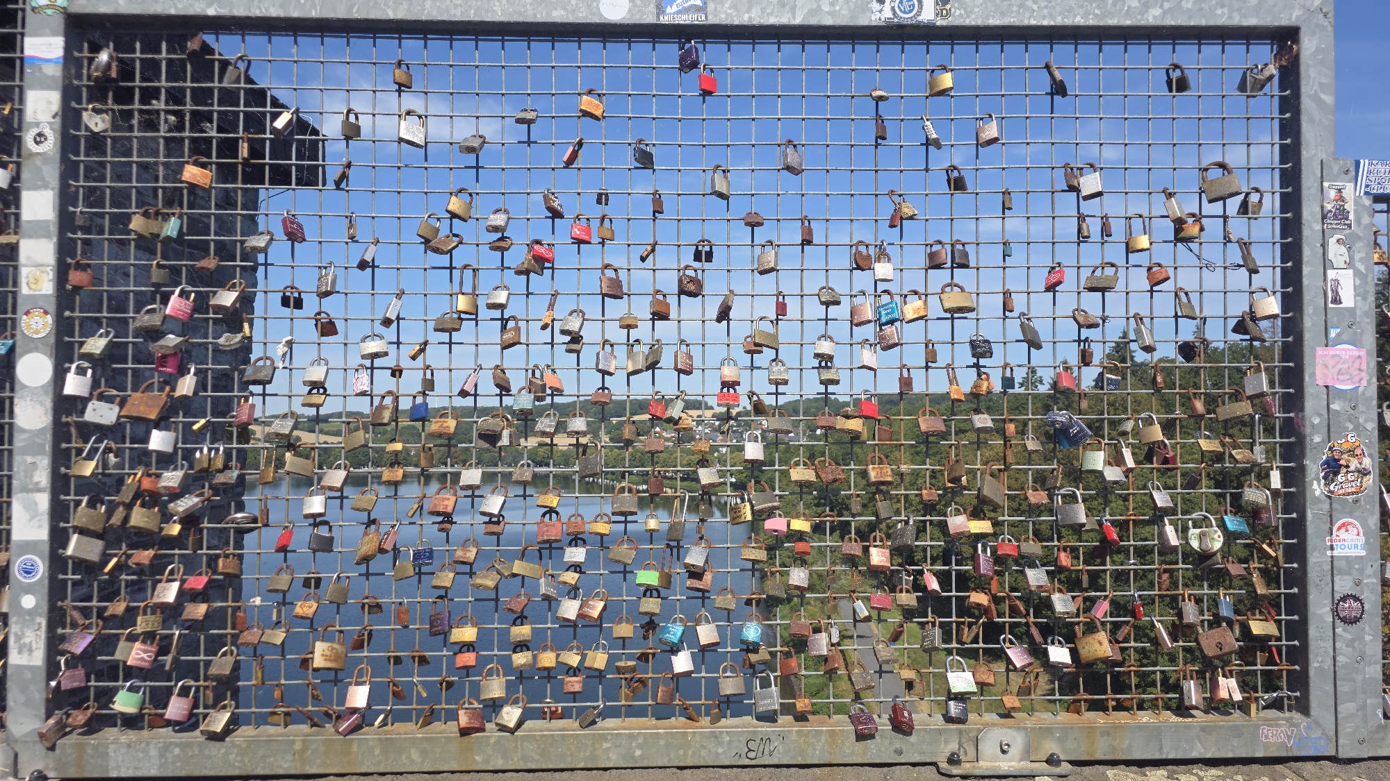 This image shows a metal fence covered in a multitude of padlocks of various colors and sizes, some of which are old and rusted. The fence is positioned in front of a body of water, likely a lake, with a tree-covered landscape visible on the opposite shore. The sky is blue with some clouds. The location is identified as the Möhnesee dam in Möhnesee, Germany. The padlocks are a common symbol of love and commitment, with couples often attaching them to bridges and fences as a declaration of their enduring affection. The scene is bathed in sunlight, casting shadows and highlighting the textures of the padlocks and the metal fence. The overall impression is one of a romantic and picturesque setting, with the dam serving as a backdrop for countless declarations of love.