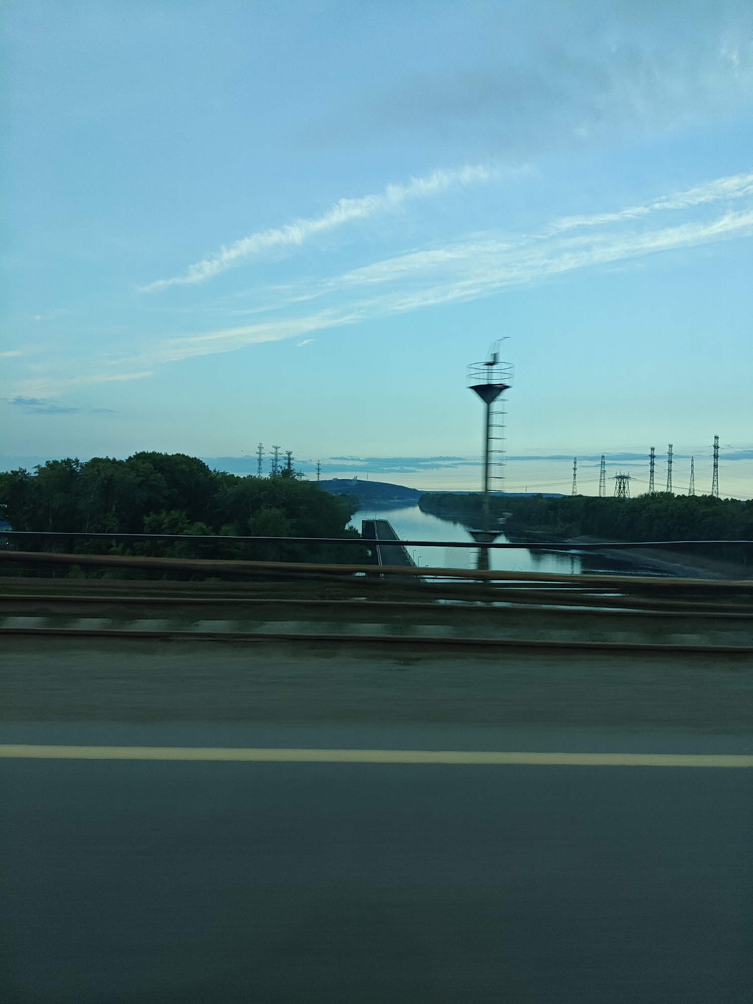 A wide-angle landscape photograph, captured from a moving vehicle on what appears to be a bridge or overpass, depicts a river scene under a clear, pale blue sky.
In the immediate foreground, a dark, somewhat reflective surface, possibly a wet road or bridge deck, occupies the bottom quarter of the frame. A single, light-colored horizontal line, likely a road marking, runs across it. Above this, several blurred, dark, parallel horizontal lines suggest railings or a protective barrier, framing the view.
The midground features a broad river or waterway extending from left to right. Along the far bank, a dense expanse of dark green trees forms a natural boundary. Beyond the treeline, a series of tall electrical transmission towers (pylons) with visible power lines are scattered across the landscape, indicating industrial development. A prominent, tall, slender structure with a circular platform at its top stands in or near the river, exhibiting significant motion blur due to the photographer's movement. This structure could be a navigational aid, an observation tower, or part of a facility.
The background shows distant landforms or hills under a vast, light blue sky adorned with scattered, wispy white clouds. The soft, even lighting suggests the time of day is either early morning or late afternoon. No people are visible in the image, and there is no discernible text. The scene blends natural riverine environment with human-made infrastructure, consistent with a location such as Chaikovsky, Russia, on the Kama River.