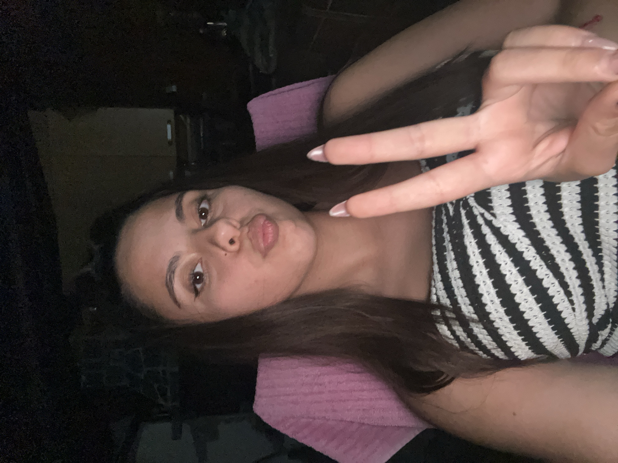 A medium close-up image depicts a female individual with long, dark hair and a medium complexion, photographed in a dimly lit environment in Unknown, Unknown. She has dark eyes, wears visible eyeliner, and exhibits a puckered lip expression. Her right hand is raised, forming a peace sign with extended index and middle fingers. Her fingernails are long and coated with a light, possibly iridescent, polish. She is attired in a strapless top featuring a black and white diagonal stripe pattern, consistent with a crocheted or knitted texture. A small, dark mole is visible on her left upper arm. Behind her, a pink, textured fabric object, likely a towel draped over a chair, is visible on the left side of the frame. The background is largely dark and indistinct, with faint structural outlines suggesting an indoor setting or a sheltered outdoor area at night.