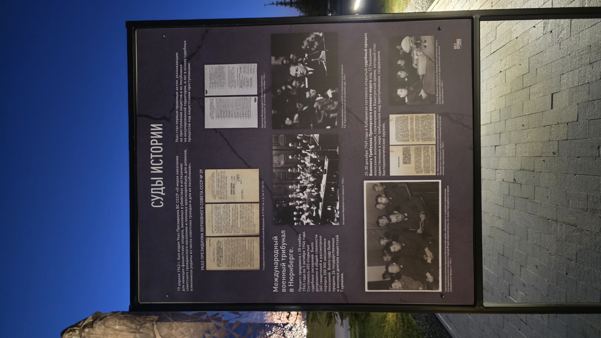 Here is a description of the image:
**Scene:** The image shows an informational display panel, possibly part of an outdoor exhibit. The panel is black with white and black text and historical black and white photographs. The panel’s subject matter appears to be about war crimes trials, specifically the Nuremberg trials and trials in Khabarovsk, Russia.
**Text (translated from Russian):** The main title is "COURTS OF HISTORY”. The text below the heading references a decree from April 1943 concerning punishment for Nazi-Fascist criminals, spies, and traitors. It further describes this decree as a foundational legal act for trials against Nazi criminals. The panel also mentions the International Military Tribunal in Nuremberg, and that the trials occurred from November 20, 1945, to October 1, 1946. It states that there were 403 open court sessions with 360 witnesses examined. The panel indicates the trials of former members of the Kwantung Army in Khabarovsk in December 1949.
**Images:** The images on the panel include photographs of legal documents, images of the Nuremberg trials, and photographs of individuals accused of war crimes during the Khabarovsk trials.
**Environment:** The panel is outdoors, with a dark blue sky in the background. There is a stone statue or monument and some greenery visible below the display. The ground nearby is paved.
Based on the identified location in the city of Zaitsevo (Verevskoe S/P), Russia, this display may be a local exhibit or memorial related to World War II and its aftermath.
