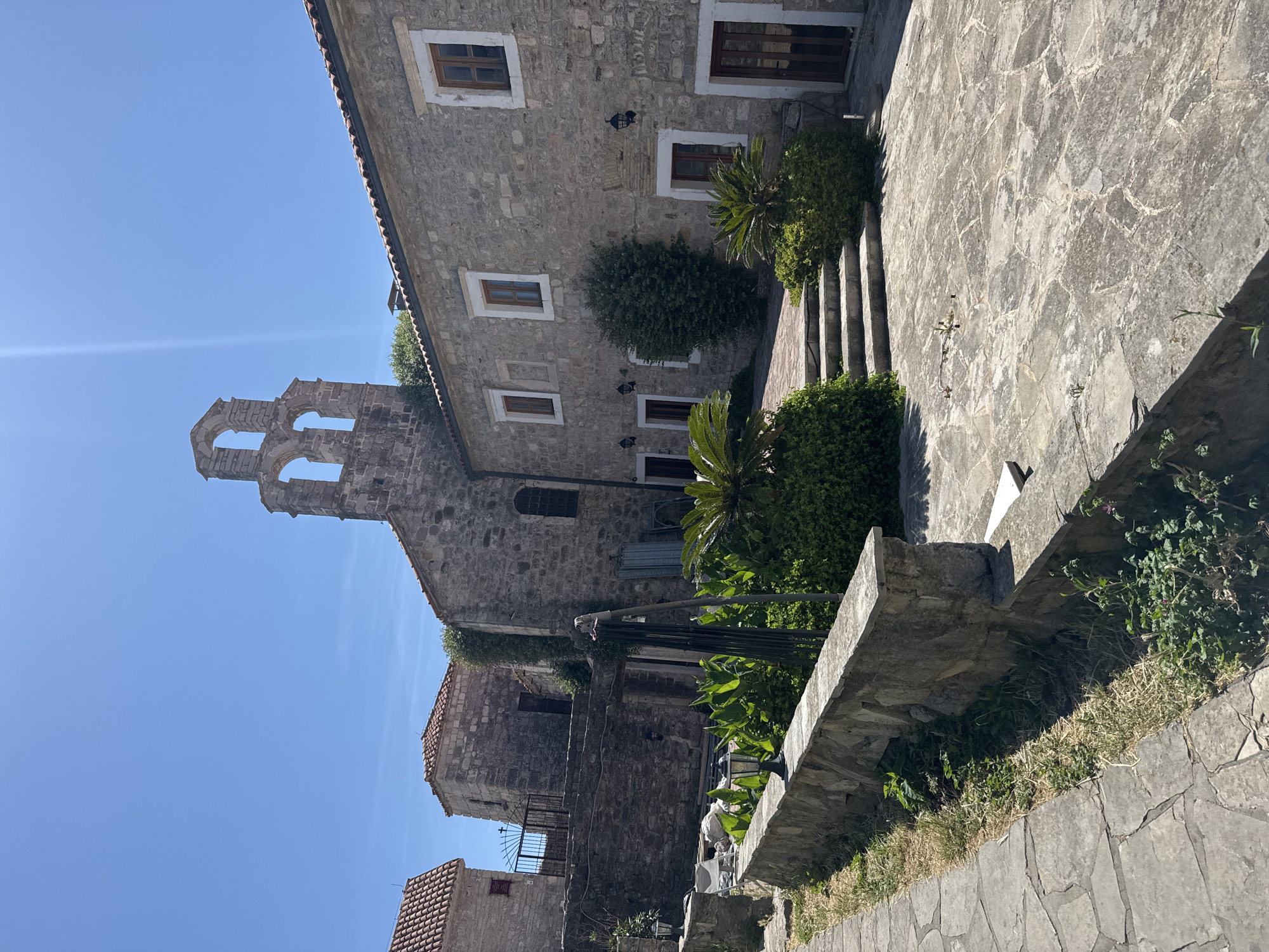 The image depicts an exterior view within the Old Town of Budva, Montenegro, under a clear, bright blue sky. The scene is dominated by traditional stone architecture and Mediterranean flora, illuminated by strong sunlight causing distinct shadows and a visible lens flare in the upper right.
In the foreground, an irregularly paved stone pathway runs along the left edge, bordered by a low, flat-topped stone retaining wall. Along this wall, two dark-colored outdoor lanterns are visible. Behind the wall, a landscaped garden area contains various green plants, including large-leafed vegetation and two small, palm-like shrubs. A dark, upright metal structure is positioned within this garden space. Further back and to the left, behind an additional stone wall, a white plastic chair is visible alongside some indistinct items.
The midground features a prominent multi-story building constructed of light-grey stone with a red-tiled roof. Its facade displays multiple rectangular windows with white frames and dark wooden sashes, along with several dark wooden doors. Black wall-mounted lanterns are affixed to this building. A set of irregular stone steps leads up to a paved courtyard in front of this structure.
In the background, additional stone buildings with red-tiled roofs are present. A distinctive stone bell tower, featuring three arched openings, rises above one of the structures on the left side. A black metal gate is visible between two buildings in the distant background. No individuals are present in the depicted scene.