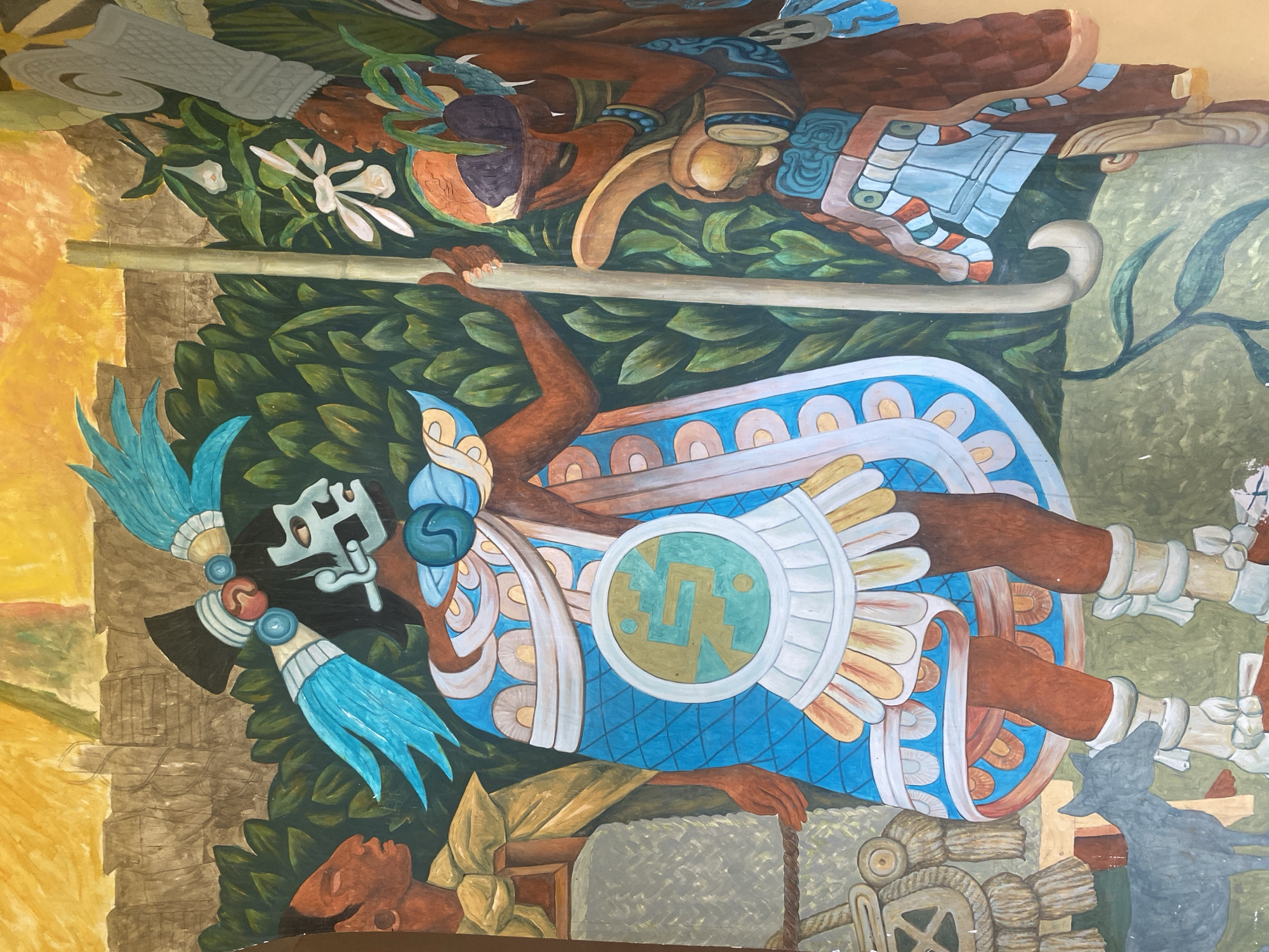 This is a close-up shot of a mural depicting figures in traditional Mesoamerican attire, set against a backdrop of lush green foliage and a textured, yellowish-brown sky. The central figure is adorned with a striking turquoise headdress with feather-like plumes and a stylized mask. They wear a vibrant blue cape decorated with a circular emblem featuring a turquoise and gold serpent-like motif, over a white and gold skirt. A turquoise sphere is visible near the figure's shoulder. To the left, another figure, with reddish-brown skin, wears a golden garment and a textured grey skirt. A grey dog stands at the lower left. To the right, a third figure, also with reddish-brown skin, holds a staff and wears an elaborate headdress. This figure carries a bowl containing dark purple and orange objects, and is adorned with turquoise bracelets and other decorative elements. The mural exhibits intricate details in the clothing, headdresses, and foliage, painted in a realistic style with bold colors. The mural is located in Kazan, Russia.