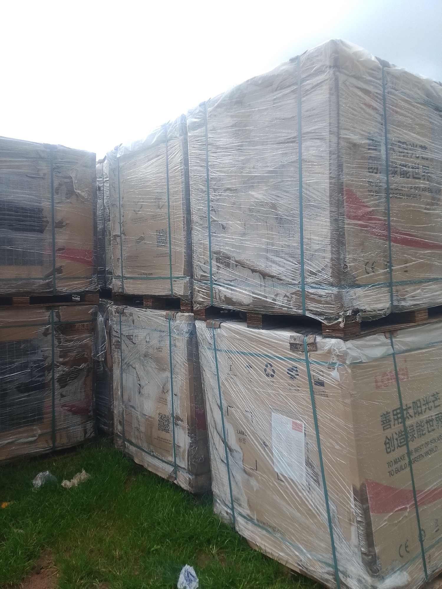 Stacked pallets of boxed items, wrapped in clear plastic, are arranged in an outdoor setting. The boxes are brown and some have writing in both Chinese and English, including "TO MAKE THE BEST OF SOLAR ENERGY TO BUILD A GREEN WORLD." The items appear to be solar panels. The background is a bright, overcast sky. The ground in the foreground is covered in green grass with some scattered debris. The location is unknown.