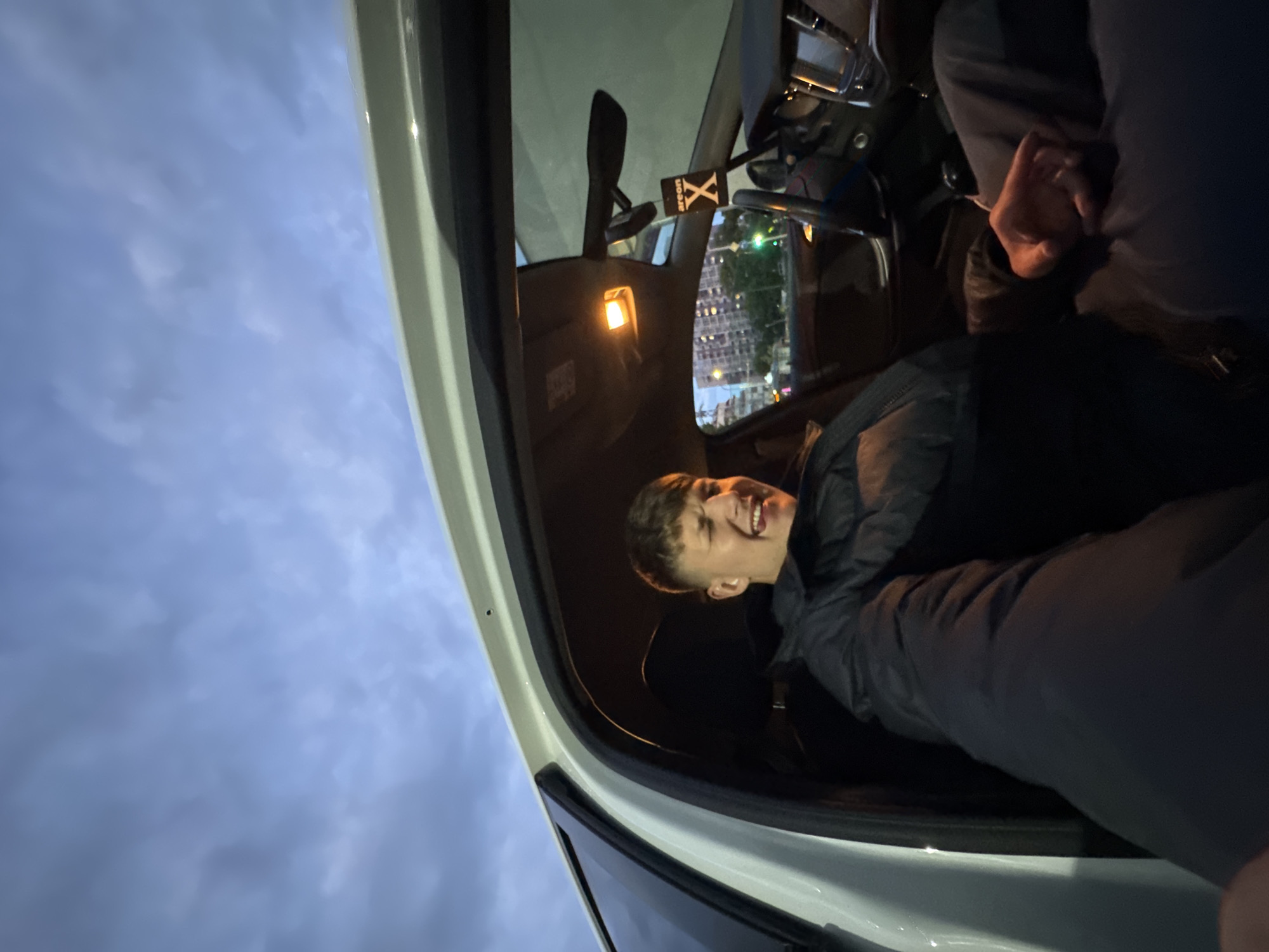 A young man is sitting in the driver's seat of a vehicle, possibly a car, with the door open. He is smiling and appears to be interacting with someone outside of the frame. The interior light of the car is on. The rear-view mirror and other internal car components are visible. Outside, a cityscape is partially visible in the reflection of the car's window, with a cluster of buildings and street lights, indicating an urban setting. The sky is overcast, suggesting it's dusk or dawn. There is an "Areon X" air freshener hanging from the rear-view mirror. While the city is not explicitly identified in the photo, the urban setting and reflection of buildings suggest that it is Burgas, Bulgaria.