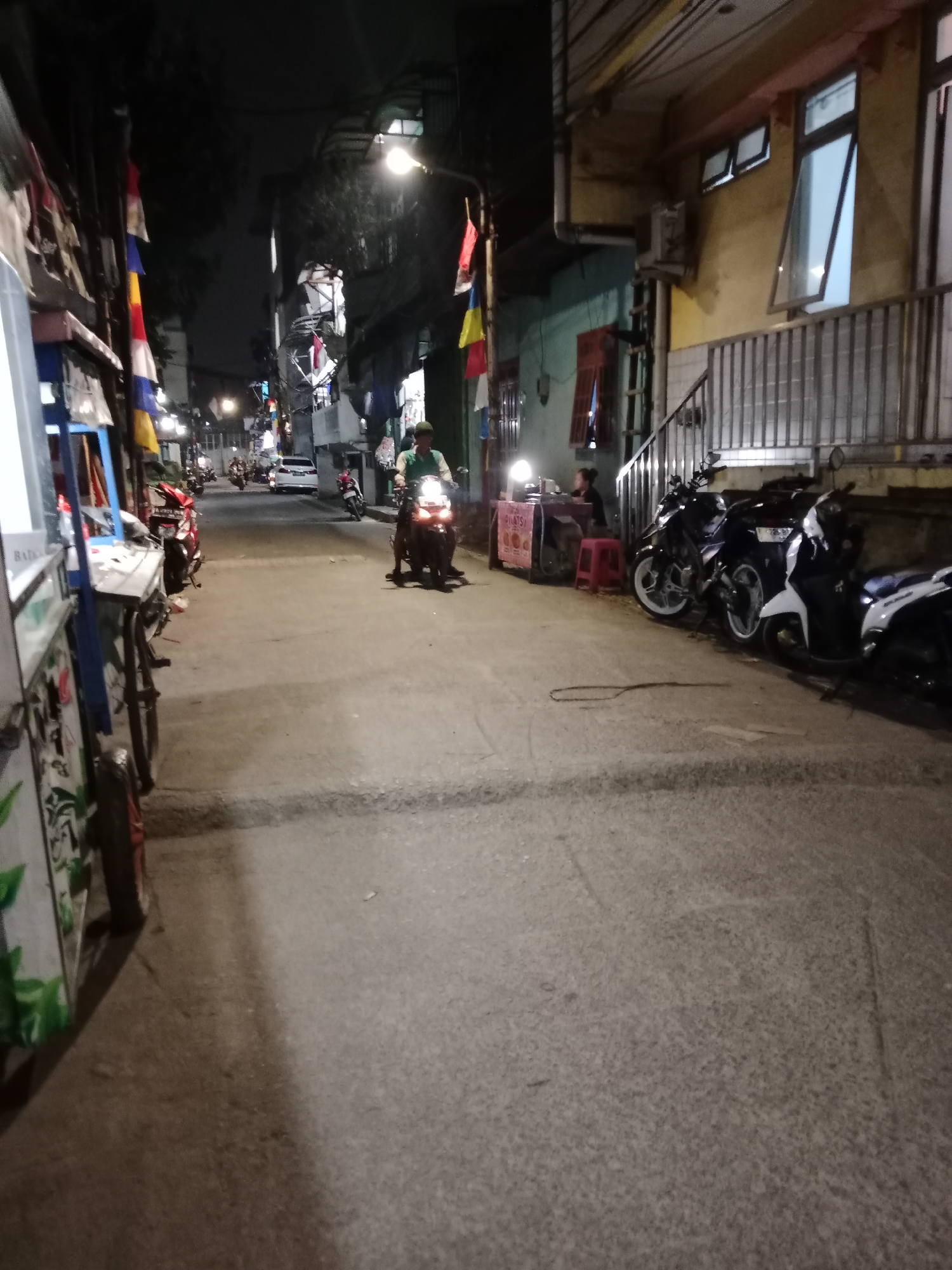 The image depicts a nighttime street scene in West Jakarta, Indonesia, likely an alley or narrow road lined with small shops and residences. The foreground is dominated by a paved road with a visible shadow cast across it. On the left side of the image, a white cart with green leaf designs is partially visible, suggesting it might be a food or merchandise stall. A dark-colored motorcycle is parked near this cart, its front wheel turned to the left.
Further down the road, a person on a motorcycle, wearing a green shirt, is illuminated by the motorcycle's headlights, suggesting they are in motion. Several other motorcycles are parked on the right side of the road. A building with yellow walls and several windows, some open, rises on the right. A metal railing runs along the front of this building. In the middle ground, a small stall with a pink counter is visible, with a person seated behind it. Red flags and other decorative elements are hung along the street, indicating a festive or public gathering. The overall lighting suggests it is evening or night, with streetlights and artificial lighting from the buildings and stalls creating illumination. The atmosphere appears to be that of a busy urban environment at night.