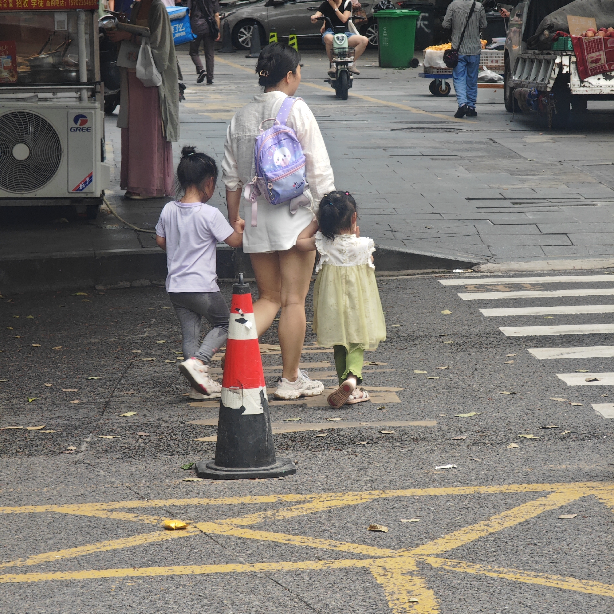 The image depicts a street scene in Yuzhong District, China. A woman wearing a white top and shorts, along with a purple backpack, is holding the hands of two young girls. One girl wears a lilac t-shirt and grey leggings, while the other wears a light green dress and green leggings. They are walking on a crosswalk, moving away from the camera. A red and white traffic cone stands nearby in the street. The pavement is asphalt with yellow crosshatch markings. In the background, there are vehicles and pedestrians, including a person on a scooter, and a food stall with a Gree air conditioner. Other commercial activities are also visible. The weather appears overcast.