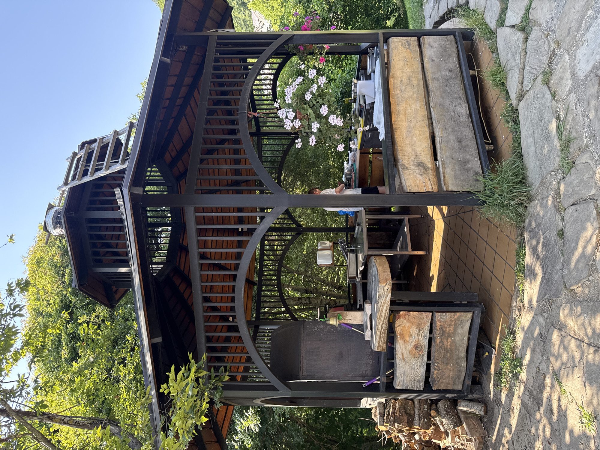 The image depicts an outdoor kitchen area, likely part of a rural or recreational establishment, set against a backdrop of lush greenery and trees under a clear blue sky. The central structure is a large, multi-tiered gazebo constructed with dark metal framing and a wooden shingled roof.
Within the gazebo, an array of kitchen equipment is visible. On the left, a substantial, dark metal barbecue or smoker stands prominently. In front of it, a rough-hewn wooden block serves as a cutting surface, supported by metal legs. Further into the gazebo, stainless steel tables are set up with various cooking utensils and containers, including what appear to be mixing bowls and a scale. A person, partially visible in a white shirt and dark pants, is attending to food preparation at one of the tables on the right side. A hanging basket filled with pink and white flowers adds a decorative touch.
The floor of the gazebo is tiled with orange square tiles. Surrounding the gazebo, the ground is covered with flagstones and patches of grass, suggesting a garden or outdoor seating area. To the left of the gazebo, a stack of firewood is neatly piled. The overall impression is that of a functional outdoor cooking space, possibly used for events or a restaurant with an emphasis on grilled or smoked foods, located in a natural, serene environment. The presence of a ladder leading to the upper level of the gazebo indicates potential for storage or access to a higher vantage point. The context of the city of Progress, Russia, is not visually evident from the image itself.