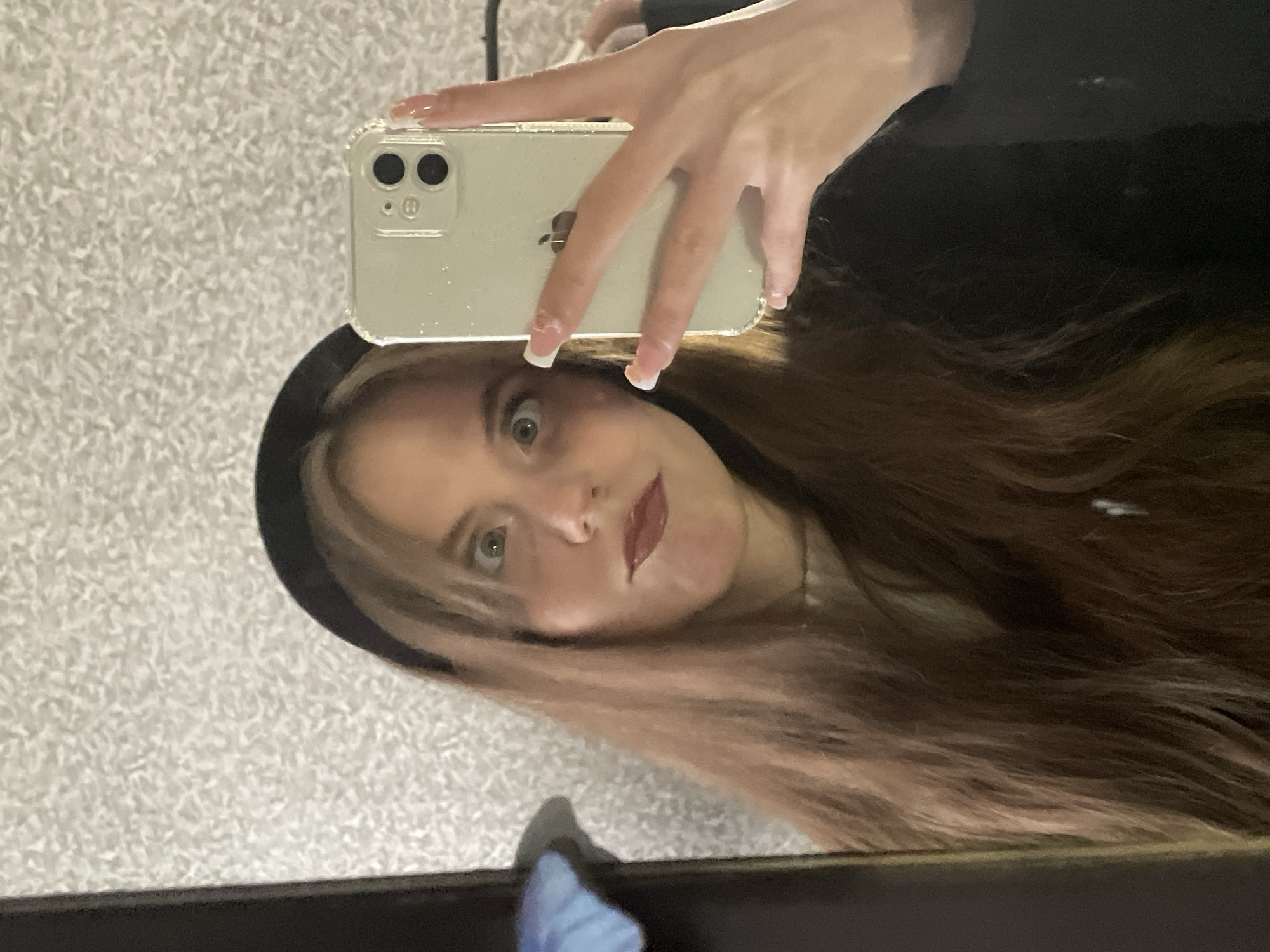 A young woman with long brown hair and a black headband takes a selfie with an iPhone. She is holding the phone in her left hand, positioned above her head. Her nails are painted with white tips. She is looking at the camera with a surprised expression. The background appears to be a textured wall. There is no evidence to suggest that this image was taken in the city of Фоки, Russia.
