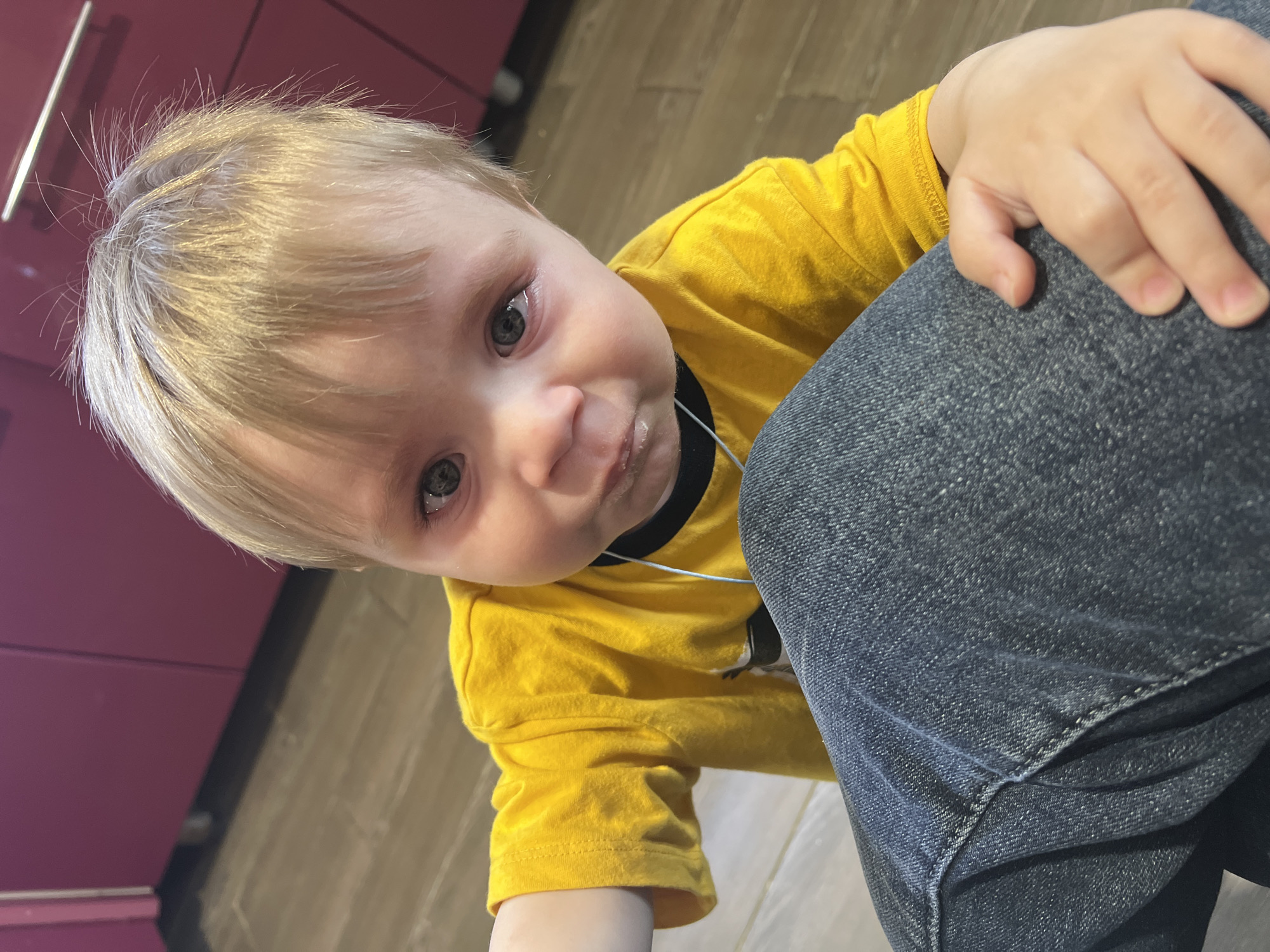 A young child with blonde hair and blue eyes wearing a yellow shirt with a black collar is looking up at the camera. The child has a small amount of liquid or residue around their mouth. Their hand is resting on a person's leg clad in blue jeans. The setting appears to be indoors with a wooden floor. A pink cabinet or piece of furniture is visible in the background. There is no indication of a city location.