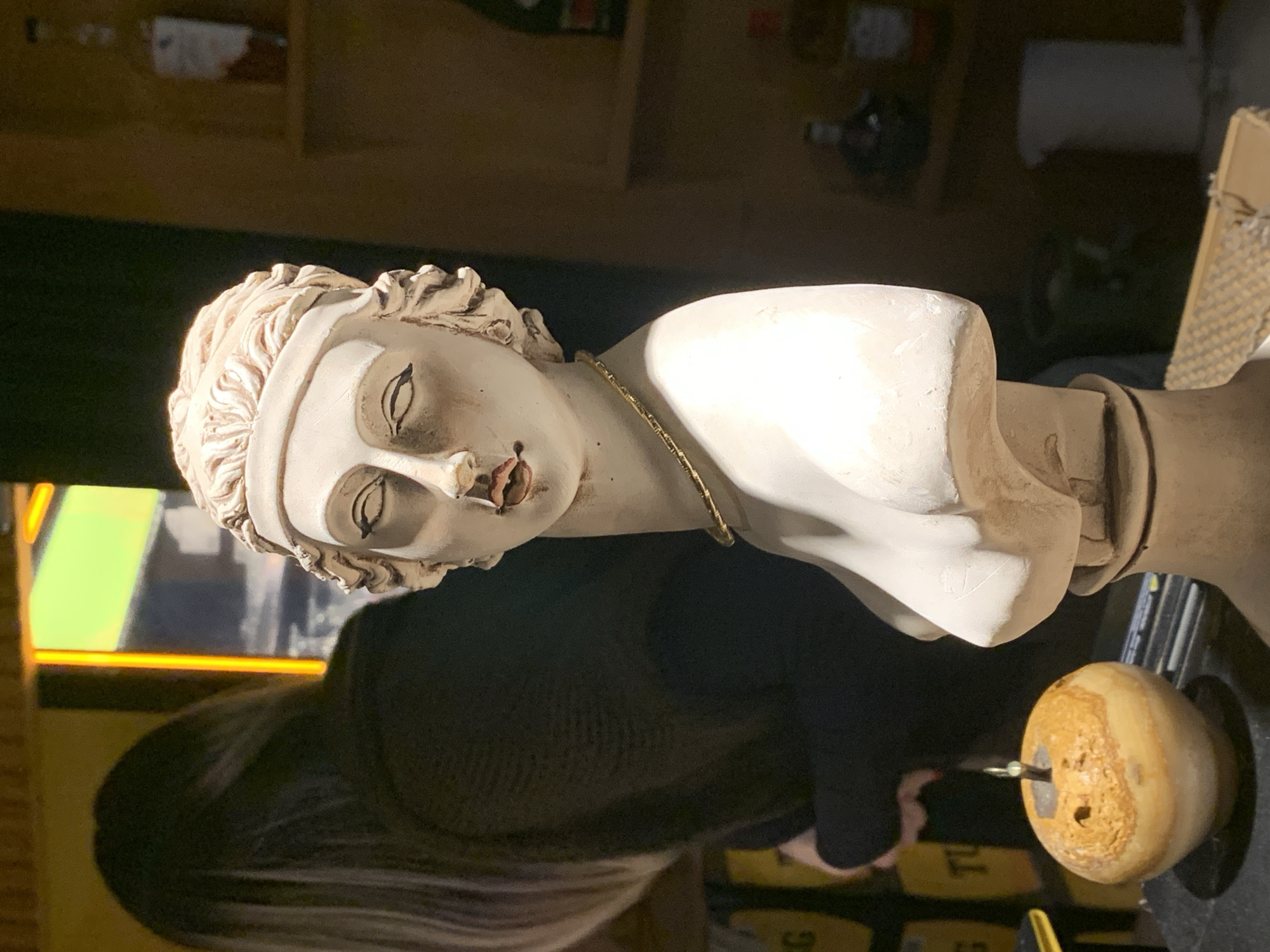 A bust of a figure with classical features and a thin golden necklace is in the foreground, situated on top of a dark-colored object with a small, yellowish-brown sphere attached. The figure's lips appear to be red. A person with long, gray hair and wearing dark clothing is visible behind the bust. A shelf with multiple bottles on it is in the background. The image may be taken in the city of Fethiye, Turkey.