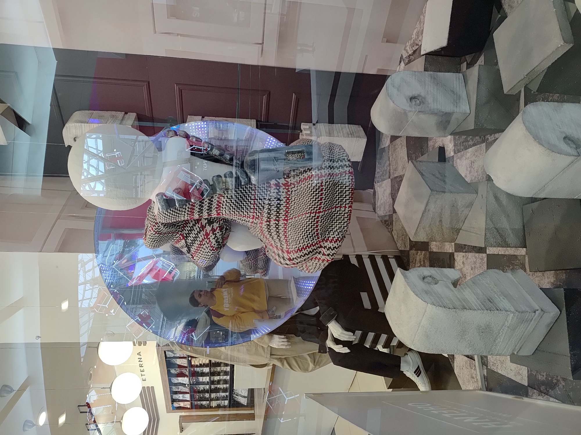 The image depicts a window display in Izhevsk, Russia, featuring mannequins dressed in contemporary clothing. One mannequin wears a yellow sweater with a burgundy collar and another is in a black suit with white stripes on the side. The display incorporates a large transparent orb or sphere around the mannequins. Overhead lighting fixtures are visible in the reflection, along with what appears to be a clothing rack in the interior of the store. The backdrop consists of geometric shapes and a tiled wall.