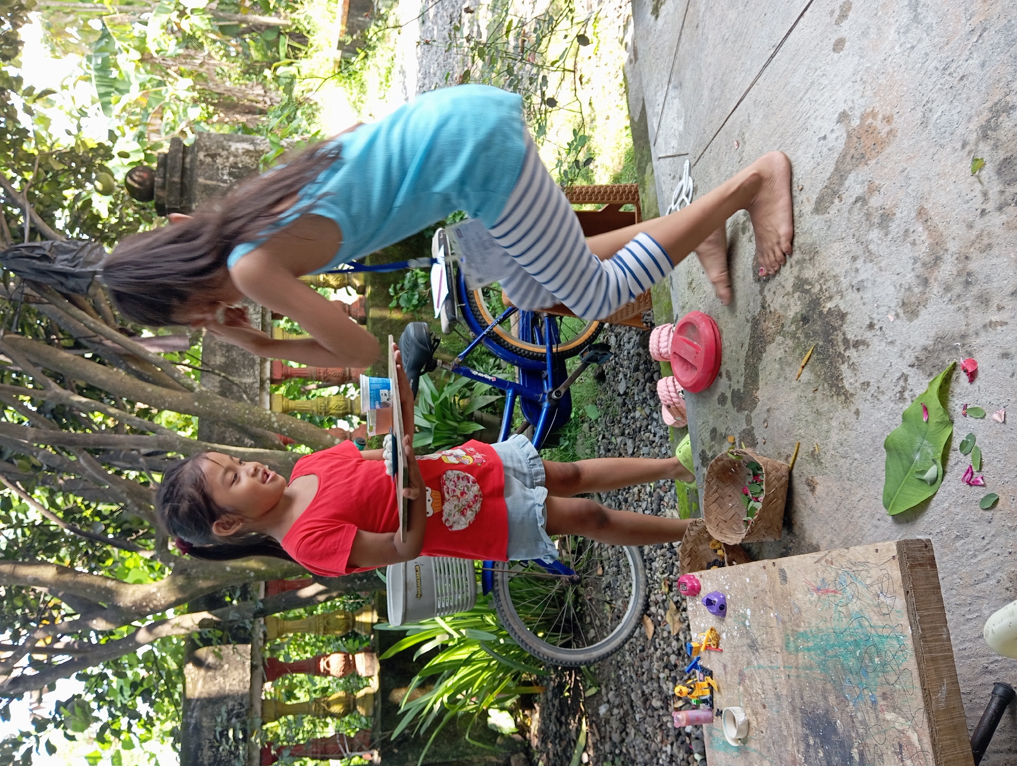 Two girls are playing outdoors in Baron, Indonesia. One girl, wearing a red shirt and blue shorts, stands holding a tablet in front of a blue bicycle. The other girl, wearing a blue shirt and striped pants, is bent over the bicycle. A wooden board with scribbles and toys is on the ground, along with a basket, sandals, and leaves. The background shows lush greenery and a weathered stone structure.
