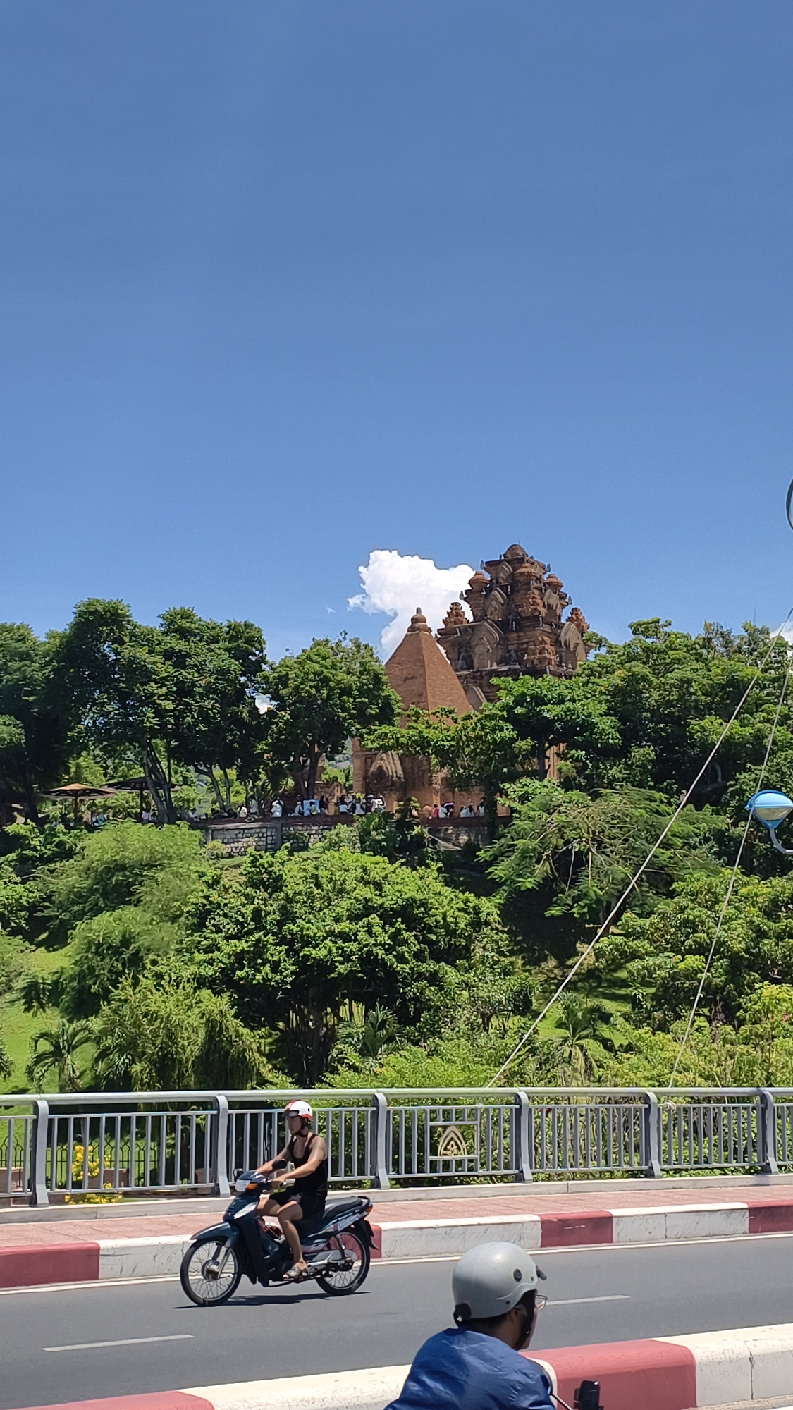 The image depicts the Po Nagar Cham Towers, a historical complex located in Nha Trang, Vietnam. In the foreground, a person rides a motorcycle on a road with a white railing to their left. The motorcycle is black and the rider is wearing a black tank top, shorts, and a white and red helmet. Behind the railing, a lush green hillside is covered with trees. Atop the hill, the brick Cham towers rise above the foliage, with a prominent pyramidal tower to the left and a more elaborate multi-tiered tower to the right. Several people are visible on the grounds around the towers, and a white cloud floats in the bright blue sky.