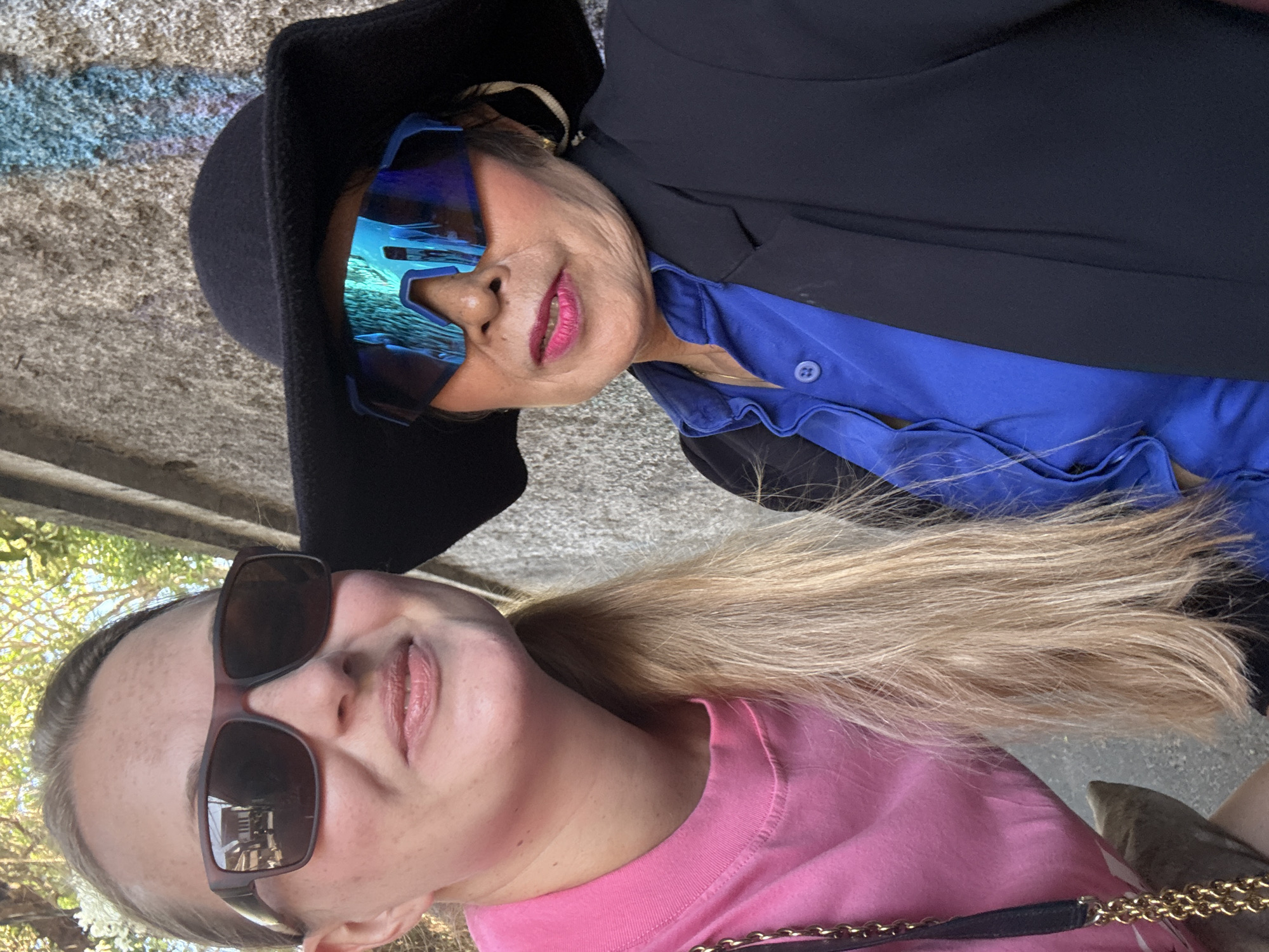 Two individuals are in the foreground, posing for a selfie against a textured, light-colored wall with some blue and pink graffiti. The person on the left, facing the camera and slightly angled, has fair skin, blonde hair pulled into a ponytail, and is wearing large, dark brown rectangular sunglasses and a bright pink long-sleeved shirt. A gold chain strap from a purse is visible across their chest. The person on the right is also facing the camera, wearing a wide-brimmed black hat, bright blue reflective sunglasses, and a royal blue collared shirt with buttons. They are also wearing a black blazer. Both individuals appear to be outdoors in a sunlit environment. The background wall suggests an urban setting, possibly a street or alleyway. The overall impression is a casual outdoor portrait.