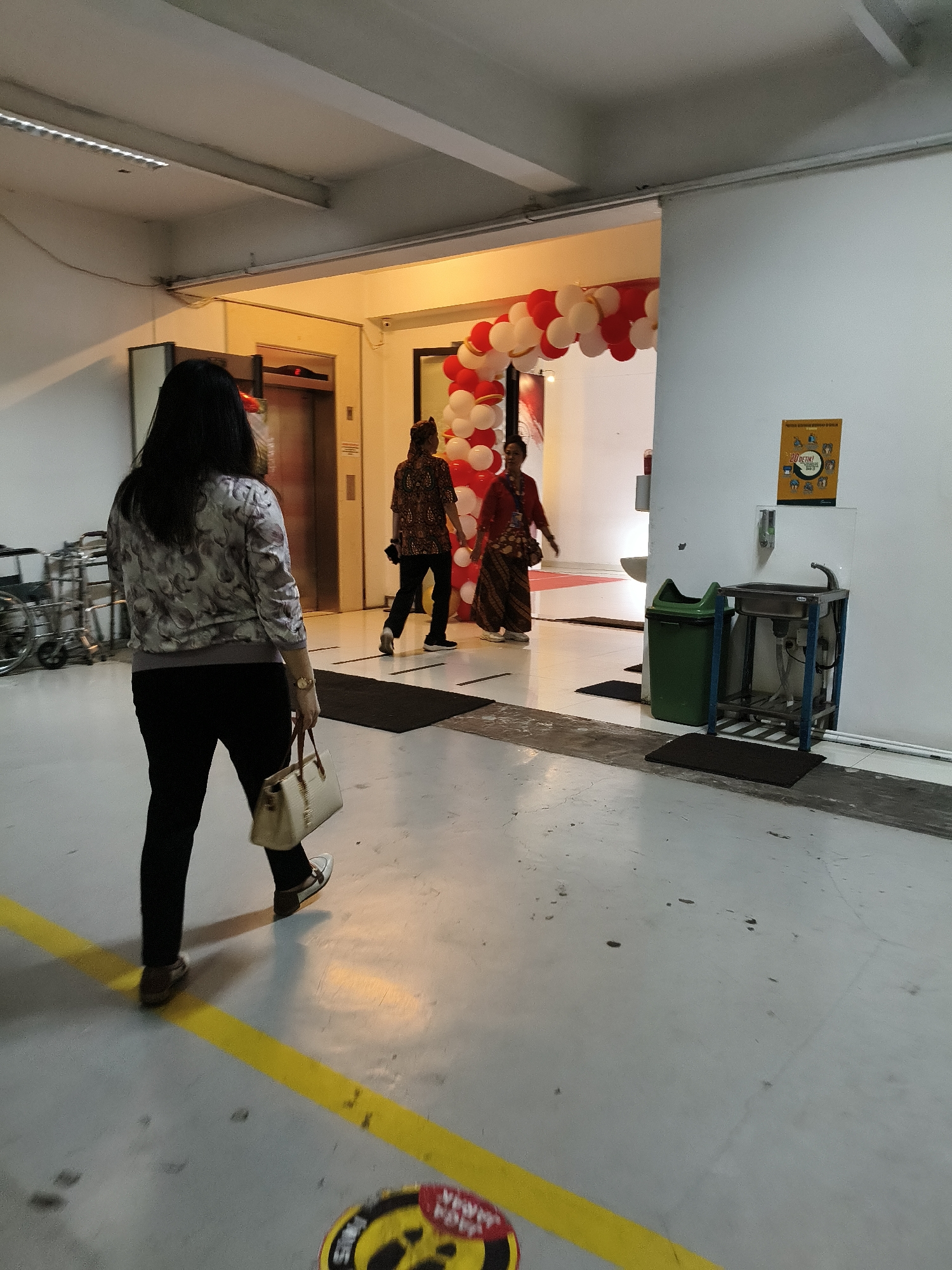 The image depicts an indoor scene with a woman walking away from the camera, carrying a light-colored handbag. She is wearing a patterned jacket over black pants and white shoes. To her right, two individuals are walking towards an archway decorated with red and white balloons. One person wears a patterned shirt and black pants, while the other is in a red top and a patterned skirt. The environment appears to be a hallway or a large room with an elevator visible in the background. A hand sanitizer dispenser and a sink are mounted on a white wall to the right. The floor is a light gray concrete with a yellow line painted on it. The overall lighting suggests an indoor commercial or public space. There is no specific indication of the city of Bandung, Indonesia in the image.