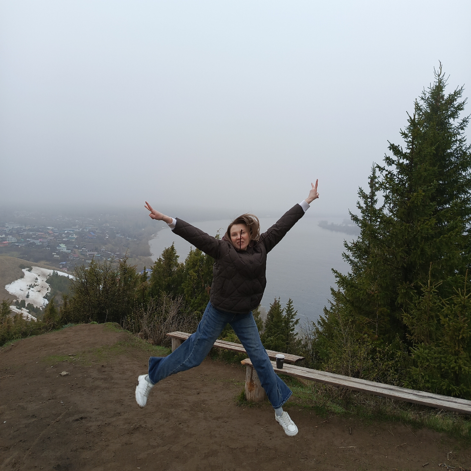 A woman jumps in the air on a hilltop in Ольховка, Russia. She has her arms outstretched making a peace sign with each hand. The woman wears a brown puffer jacket, blue jeans, and white tennis shoes. Behind her is a view of the city shrouded in fog, with a river and forest visible. To her right is a tall evergreen tree, and to her left, a wooden bench sits on the ground. The ground is dirt and grass. The sky is overcast and grey.
