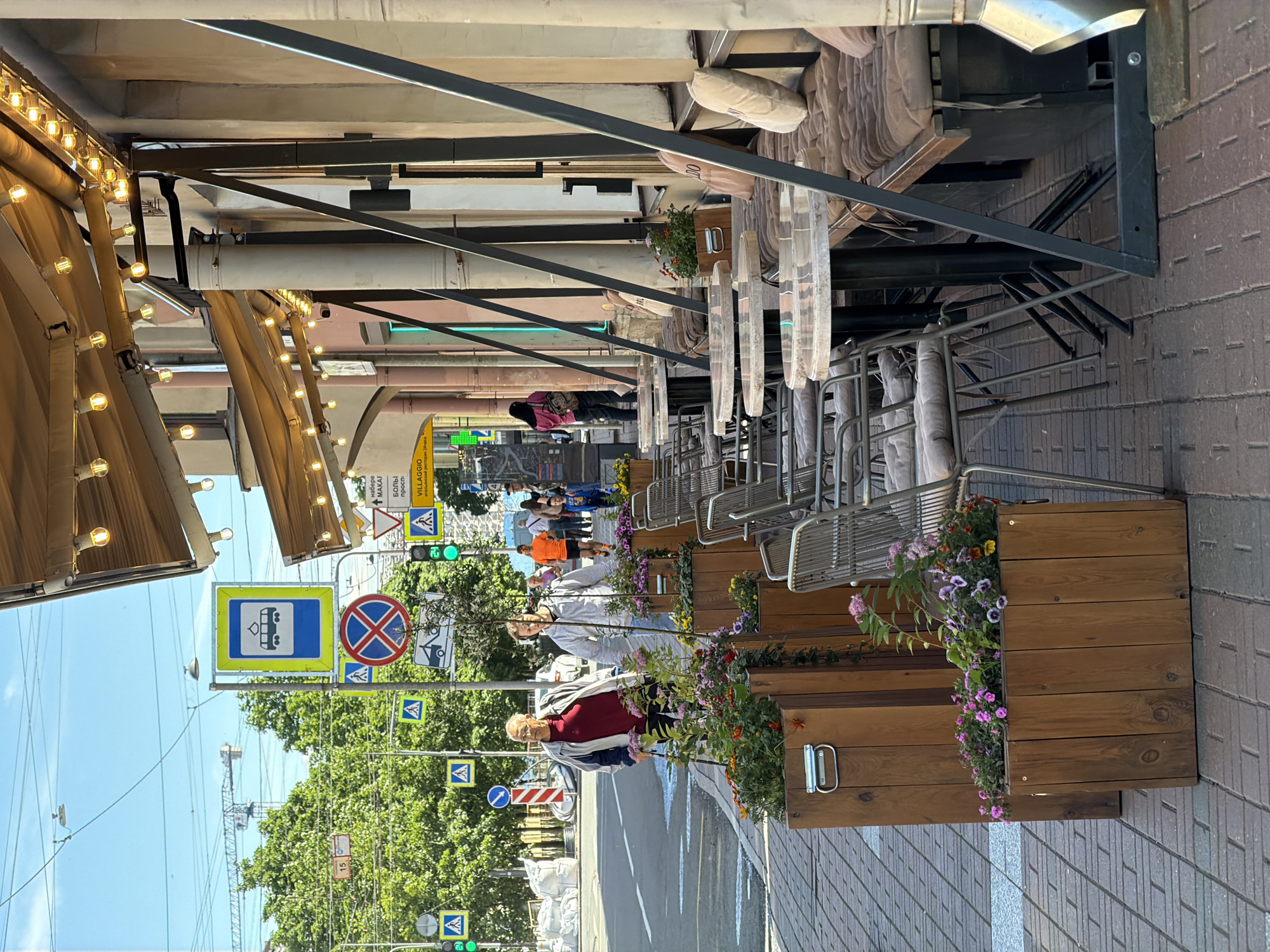 The image depicts an outdoor seating area of a cafe on a sunny day in Saint Petersburg, Russia. The cafe has a light-colored awning with string lights, and the tables are round with marble-patterned tops and black metal bases. Chairs with gray cushions are arranged around the tables. Planters with pink and purple flowers are placed along the sidewalk in front of the cafe.
In the background, there is a street with pedestrian and traffic signals. Several people are visible walking on the sidewalk and crossing the street. A tram sign is also visible. The architecture of the buildings suggests a European city, and the presence of a tram sign and the overall ambiance are consistent with Saint Petersburg.
The scene is captured from a slightly elevated angle, looking towards the cafe and the street. The focus is on the cafe's outdoor seating and the sidewalk, with the background elements providing context.
