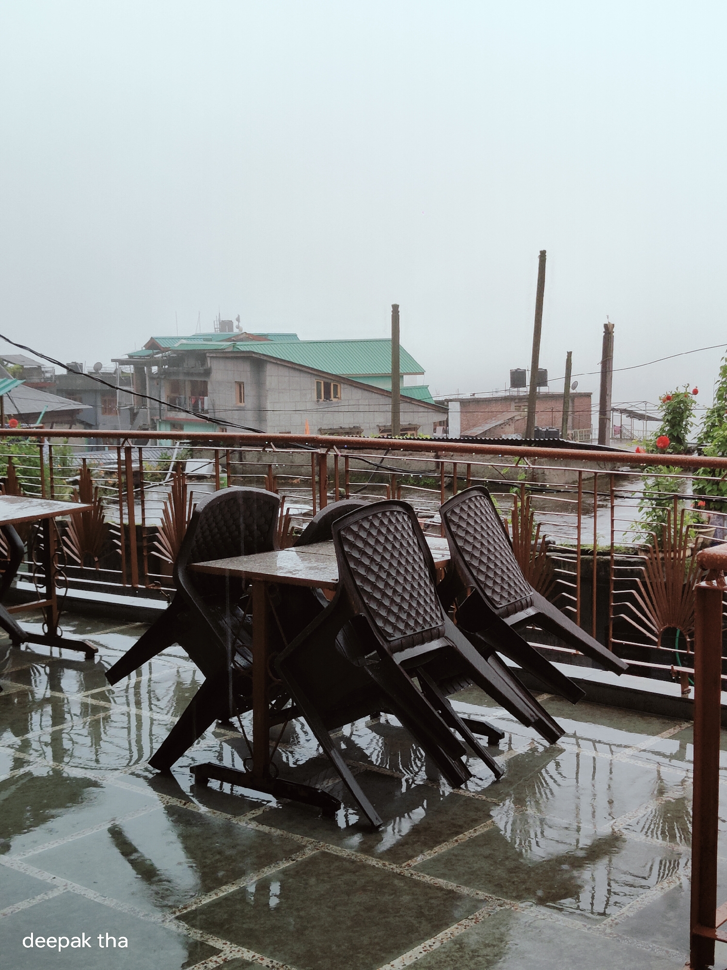 The image depicts an outdoor seating area of a cafe or restaurant in Vashisht, India, on a rainy day. Several dark plastic chairs are stacked and arranged around square tables. The ground, made of large square tiles, is wet and reflects the surrounding objects due to the rain. In the background, buildings with green roofs are visible, along with power lines and utility poles. The sky is overcast and hazy, indicating a foggy or misty weather condition typical of the region. The overall scene suggests a quiet, deserted ambiance due to the weather. The watermark "deepak tha" is present in the bottom left corner of the image.