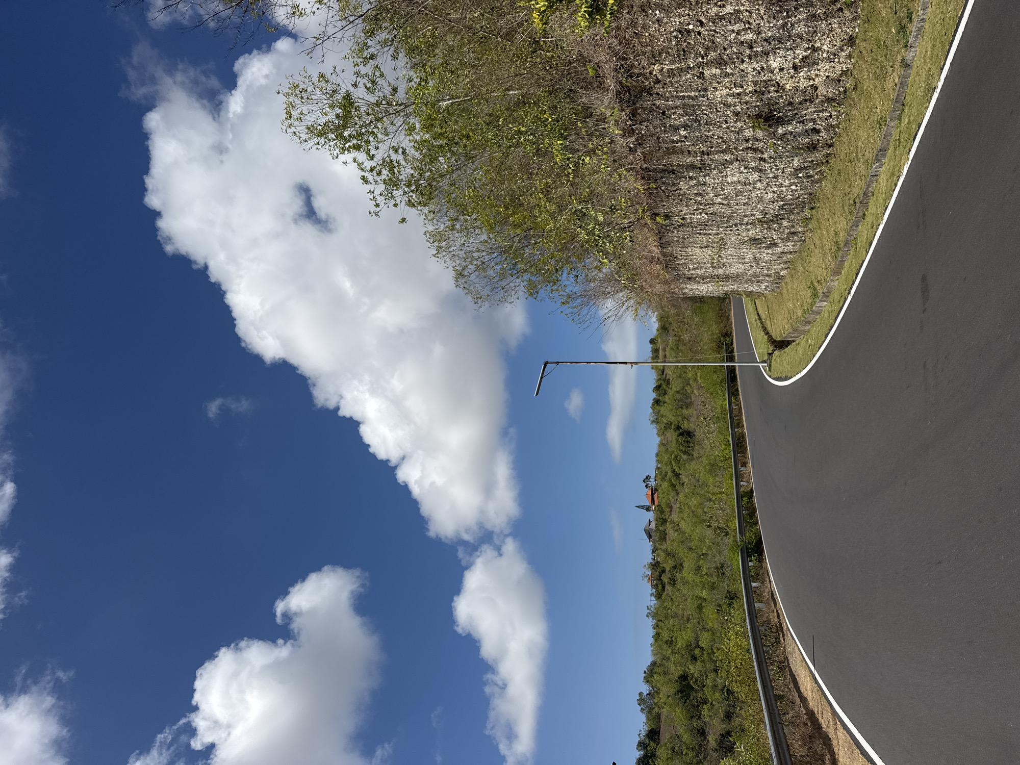A paved, winding road curves through a hilly, vegetated landscape under a bright blue sky dotted with fluffy white clouds. To the right of the road, a rough-hewn stone wall rises, topped with a dense growth of trees and shrubs. A metal guardrail runs along the left side of the road, separating it from a grassy embankment. In the distance, on a hilltop, several buildings with red-tiled roofs are visible. A modern lamppost stands near the road's edge. The scene appears to be in Kuta Selatan, Indonesia, possibly a scenic route or overlook area.