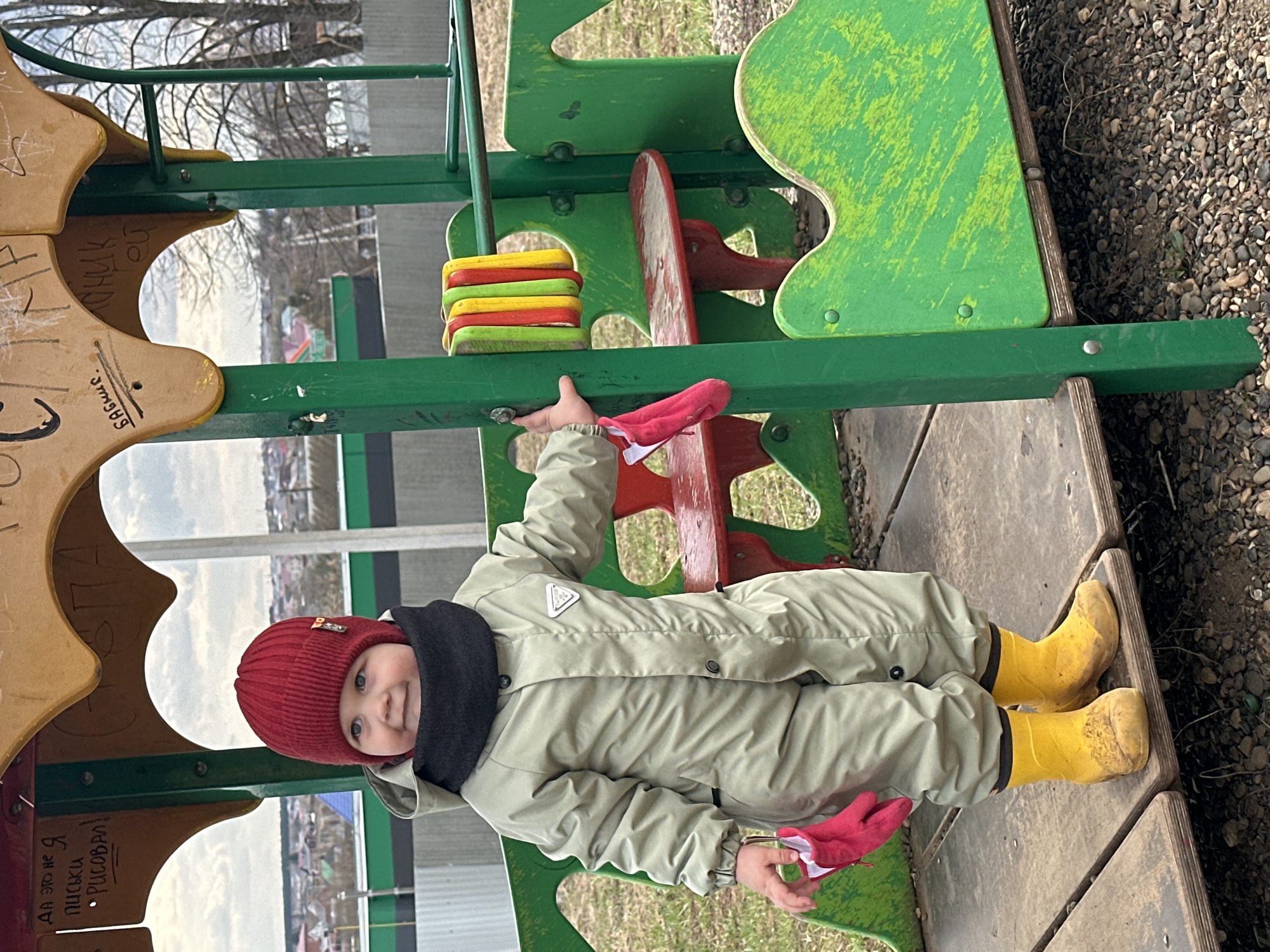 A young child wearing a red hat, dark gray scarf, light green jumpsuit, and yellow boots is playing on a playground. The child is holding onto a green metal bar attached to a playground structure. The playground structure has elements of green, red, and beige. The ground around the playground is covered in small pebbles. In the background, there are houses and trees. The location of the playground is in the city of Ludorvay, Russia.