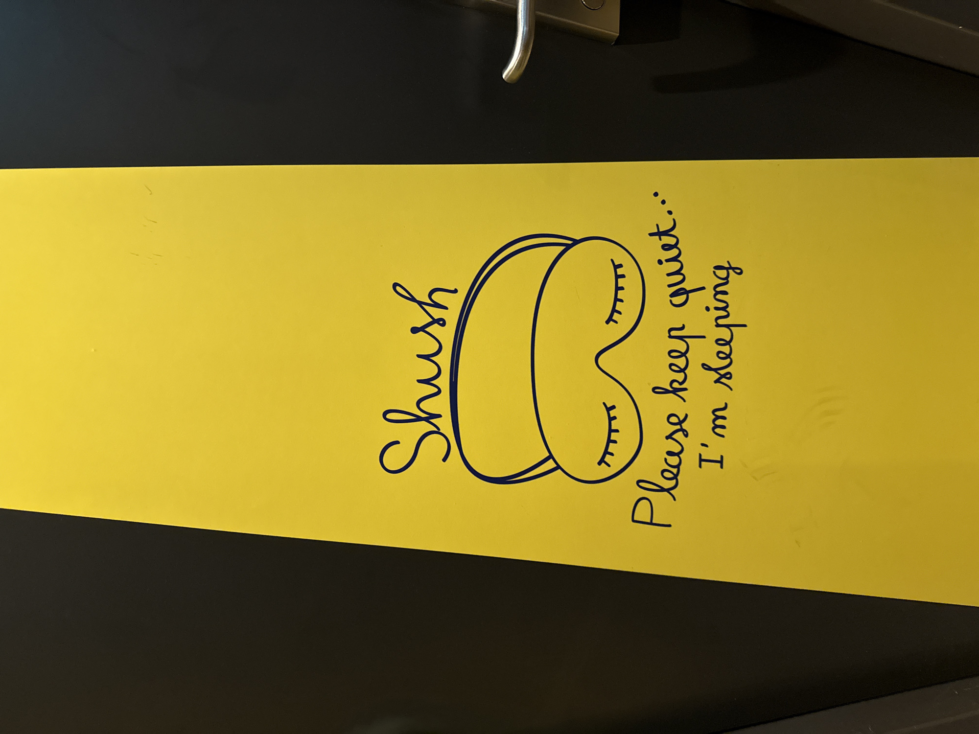 This image shows a yellow sign on a dark gray surface, possibly a door. The sign has handwritten text in dark blue ink. At the top, it says "Shush". Below that, there is a drawing of a pair of closed eyes with eyelashes, resembling an eye mask. Underneath the drawing, the text continues with "Please keep quiet..." and then "I'm sleeping". The sign is angled, suggesting it is on a vertical surface. The overall impression is a polite request for quiet. The location in Stein, Netherlands, is not discernible from the image itself. There are no people visible in the image. The background appears to be a textured, dark gray surface, with a silver door handle visible on the right side.