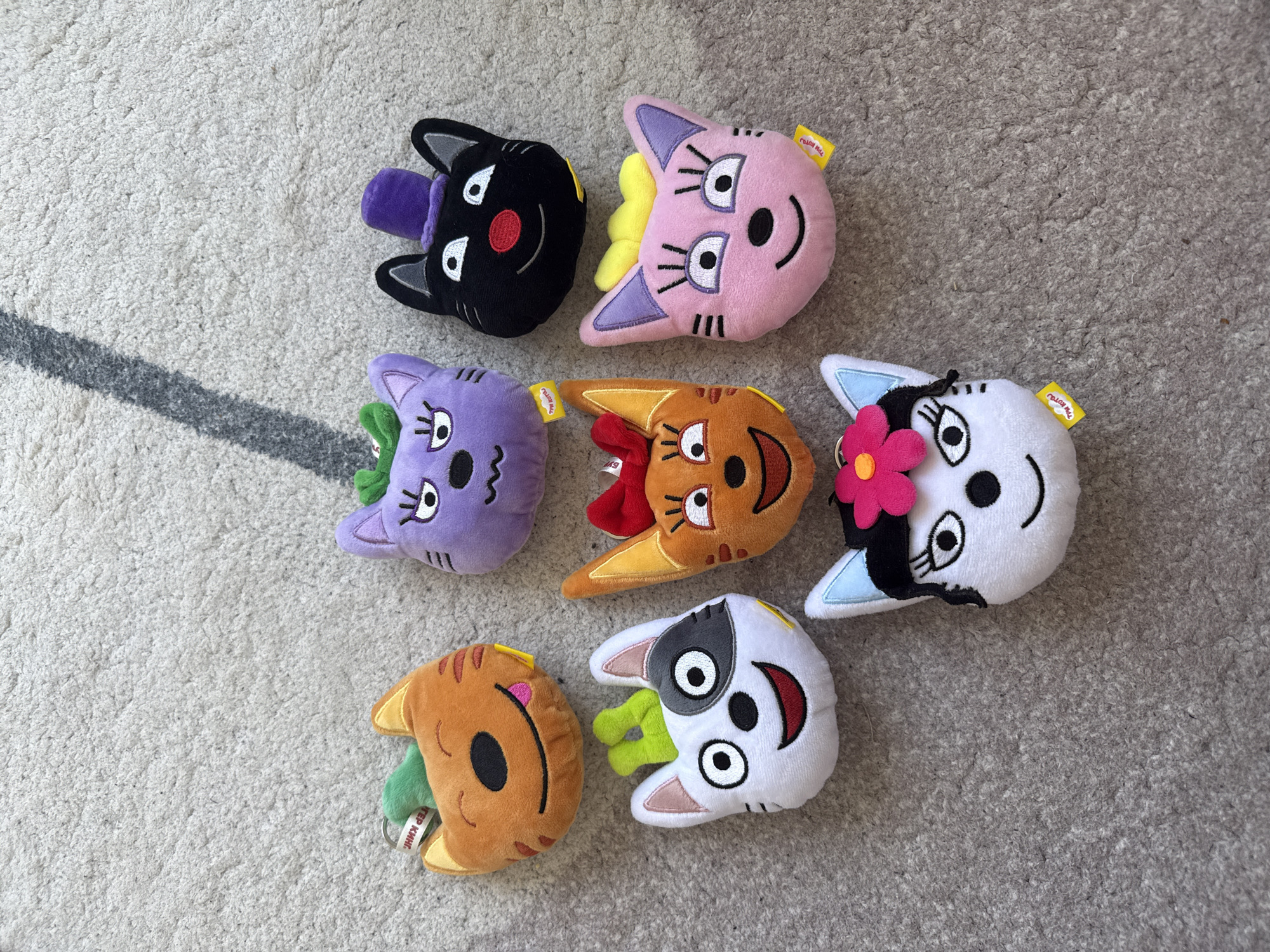 Seven cat-faced plush toys are arranged on a grey, speckled carpet. The toys are different colors and have varying facial features and accessories. The orange cat on the top left has a green stem-like object on its head and its tongue sticking out. Next to it, a purple cat has green stems on its head and a wavy mouth. The black cat in the top right wears a purple top hat and has a red nose. In the middle row, a white and black cat with green stems on its head grins widely. Next to it is an orange cat with a red bow on its head and a smiling mouth. The pink cat on the right has yellow accents on its head and a smiling mouth. At the bottom, a white cat with black hair and a pink flower on its head faces forward. All toys have small yellow tags with Russian text. The image is shot from a high angle, showing the toys from above on the carpet. There are no people present in the image, and no discernible environmental features suggest a specific location within Lyubertsy, Russia. The lighting appears to be natural, casting soft shadows.