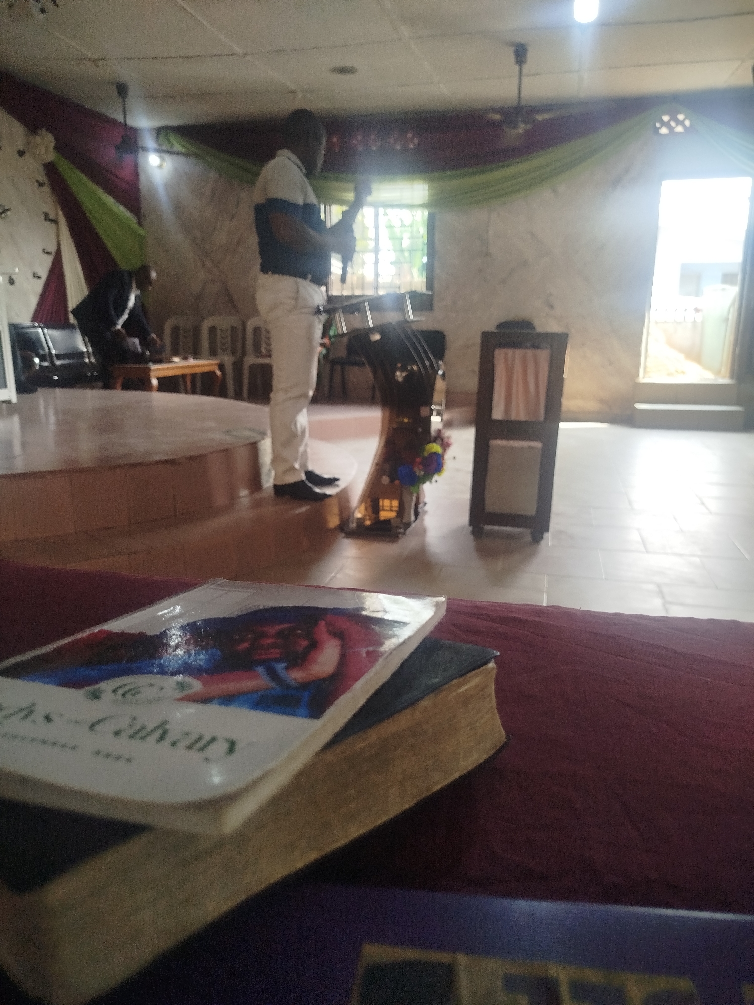The image shows the interior of a church. In the foreground, there are books stacked on a red cloth-covered surface. A man in a white shirt and pants stands near a podium decorated with flowers on a raised platform. Another man sits in the background, near chairs and a table. Decorations, including draped fabrics in green and red, adorn the background. Natural light streams through a window and an open doorway, revealing glimpses of the outdoors. The location is Unknown, Unknown.