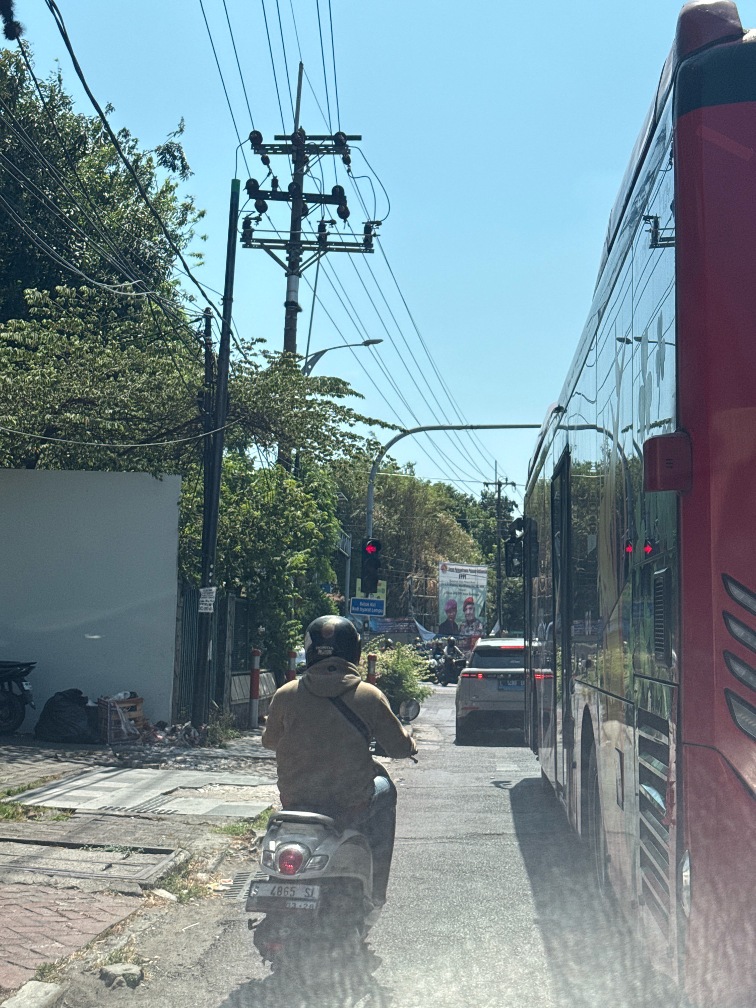 The image depicts a street scene in Surabaya, Indonesia, on a clear, sunny day. A red bus is prominent on the right side of the frame, its side reflecting the surrounding environment. A person wearing a light brown hoodie and a dark helmet rides a scooter in the foreground, heading away from the camera. The scooter has a license plate that reads "L 4865 SI". In the background, a gray SUV is visible, also in traffic. Overhead, electrical wires and a utility pole are strung across the sky. Lush green trees line the street, and a traffic light is visible, showing red. The road surface appears to be asphalt, with some debris and construction materials present on the side of the road, indicating ongoing urban activity.